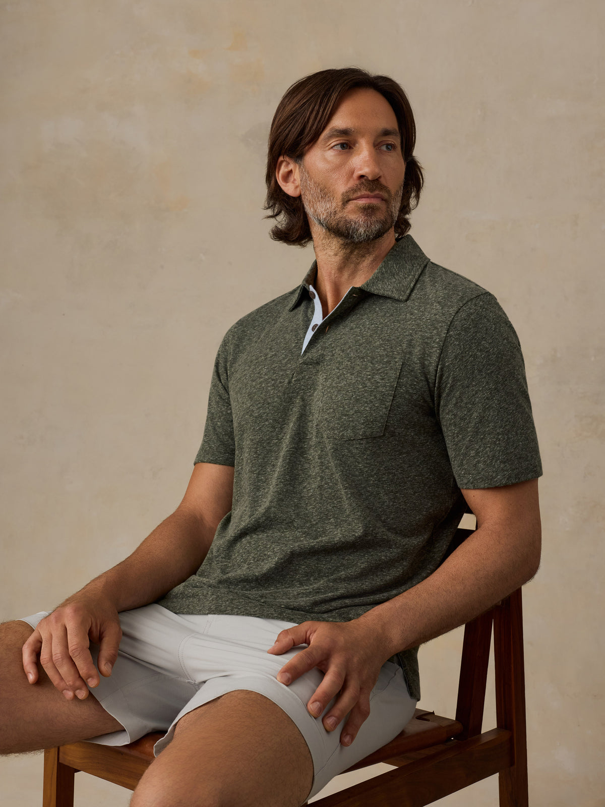 A man with shoulder-length brown hair and a beard sits on a wooden chair wearing a dark heathered green short-sleeve polo shirt and light gray shorts against a textured beige wall., image 6 of 6