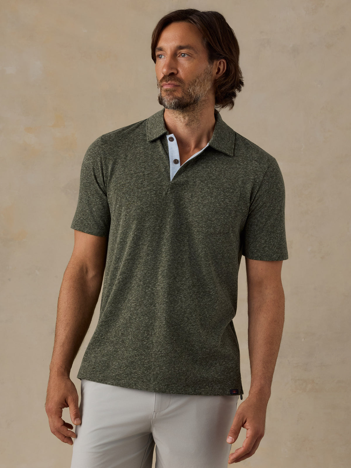 A man wearing a short-sleeve polo shirt in heathered olive green, paired with light-colored trousers, standing against a textured beige wall., image 1 of 6