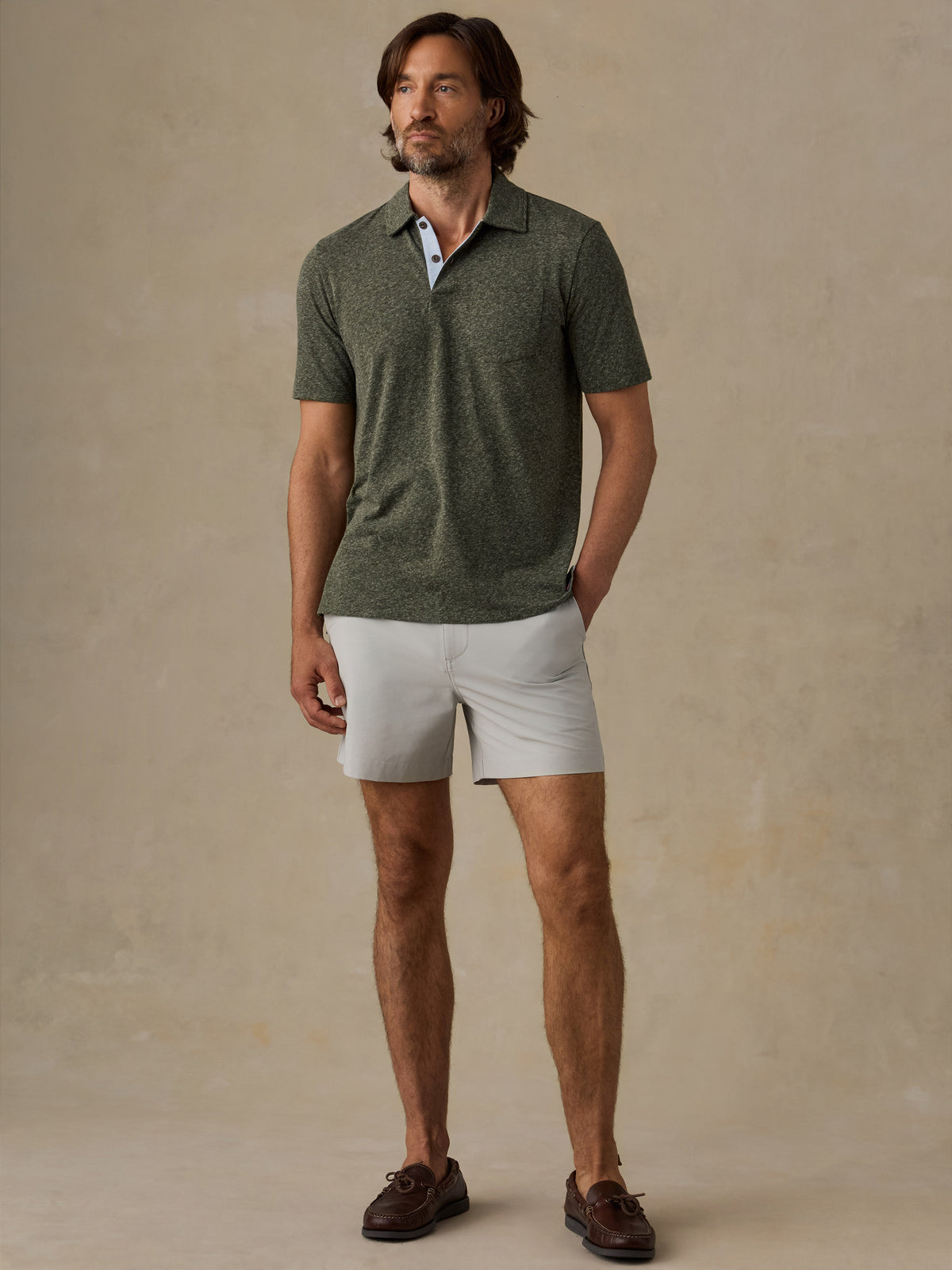A man modeling a short-sleeve heathered green polo shirt with white shorts and brown boat shoes, standing against a textured beige studio backdrop., image 4 of 6