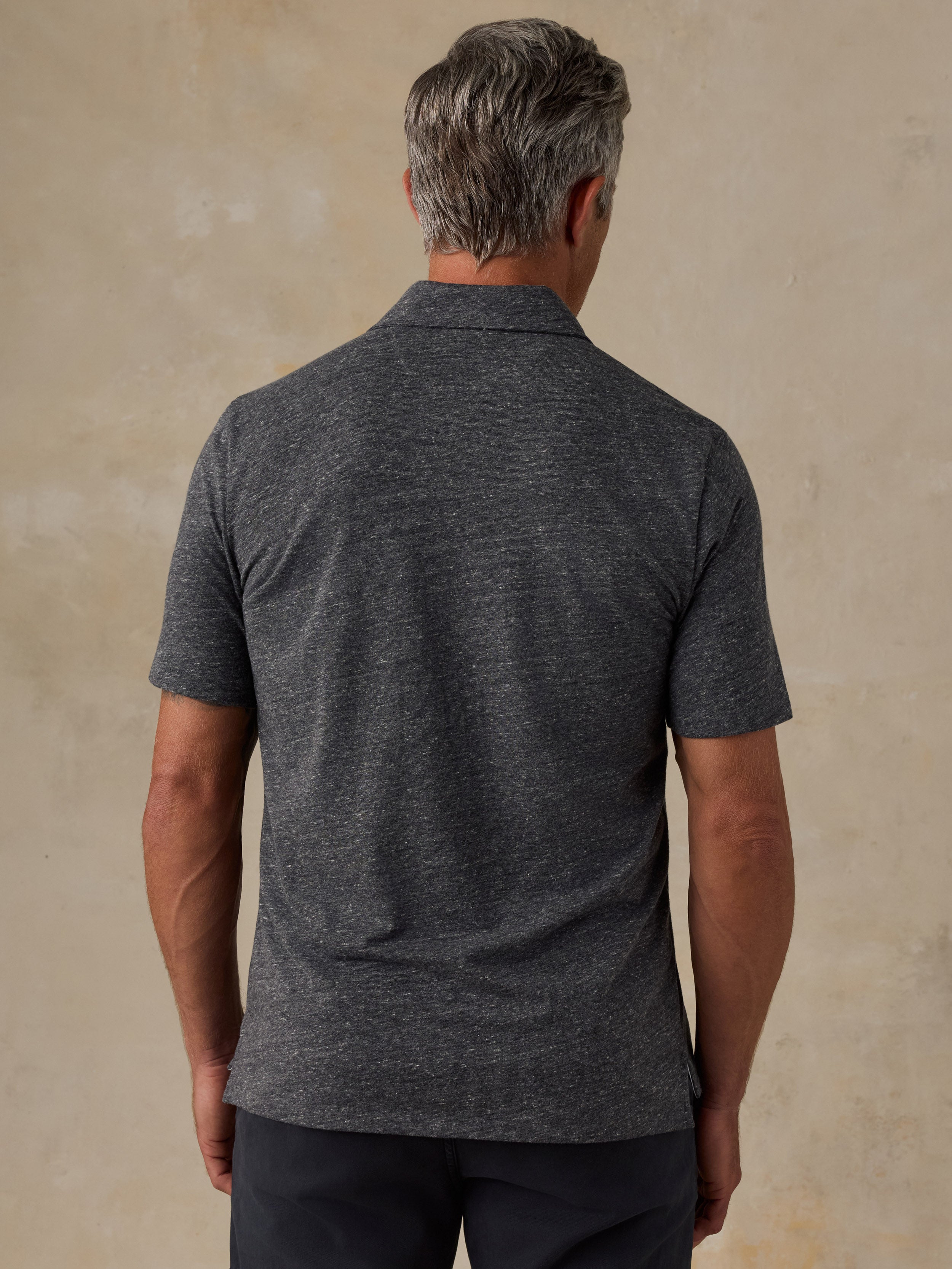 A man viewed from the back wearing a dark heather gray short-sleeve polo shirt and dark pants, standing against a textured beige wall.