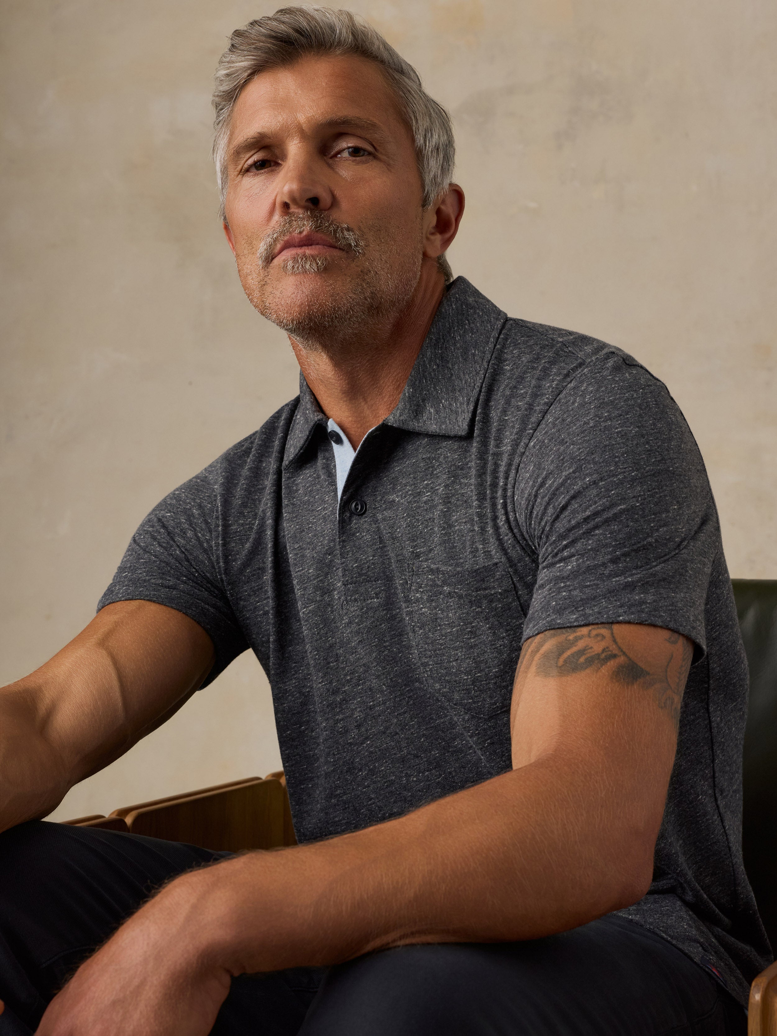 A man wearing a dark heather gray short-sleeve polo shirt, seated against a textured beige wall.