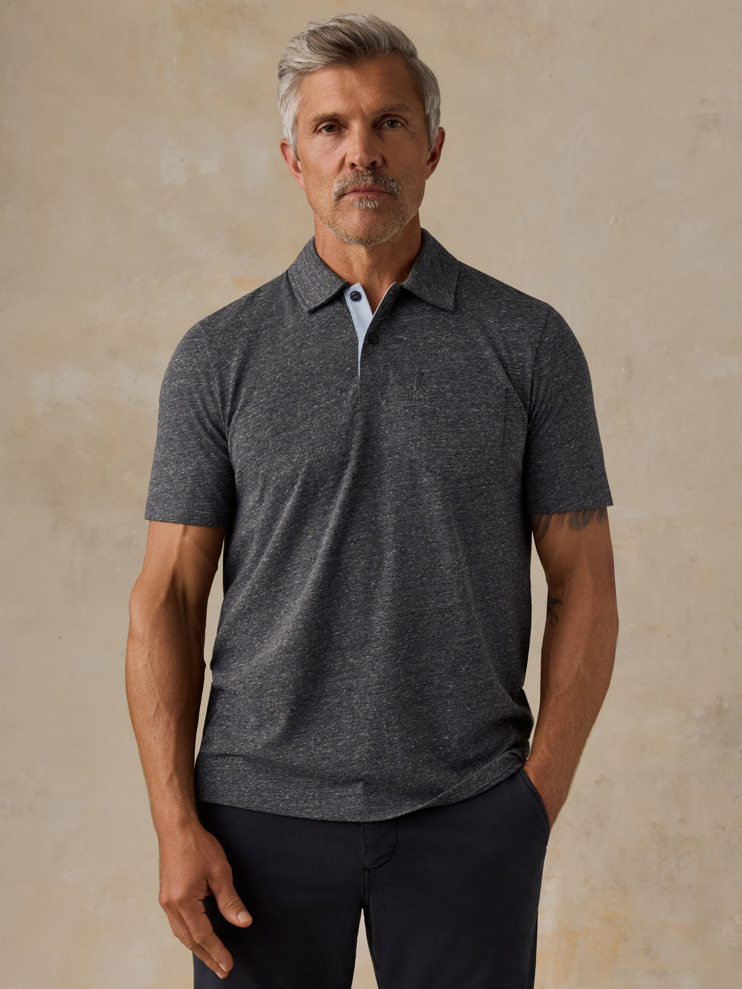 A man wearing a dark heather gray short-sleeve polo shirt with a light blue inner placket, standing against a textured beige studio backdrop.
