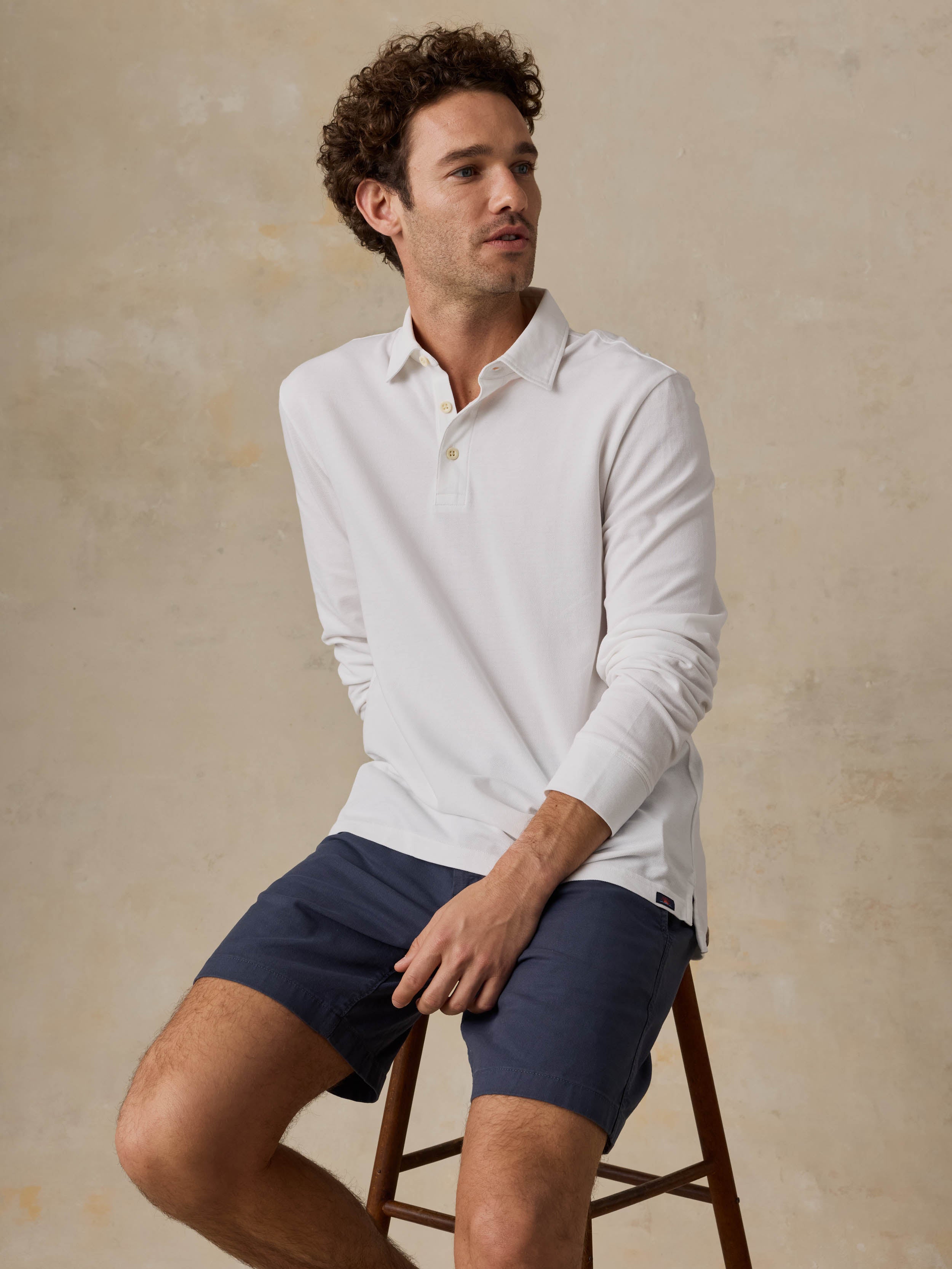 Man sitting on a wooden stool wearing a white long-sleeve polo shirt and navy shorts against a beige background