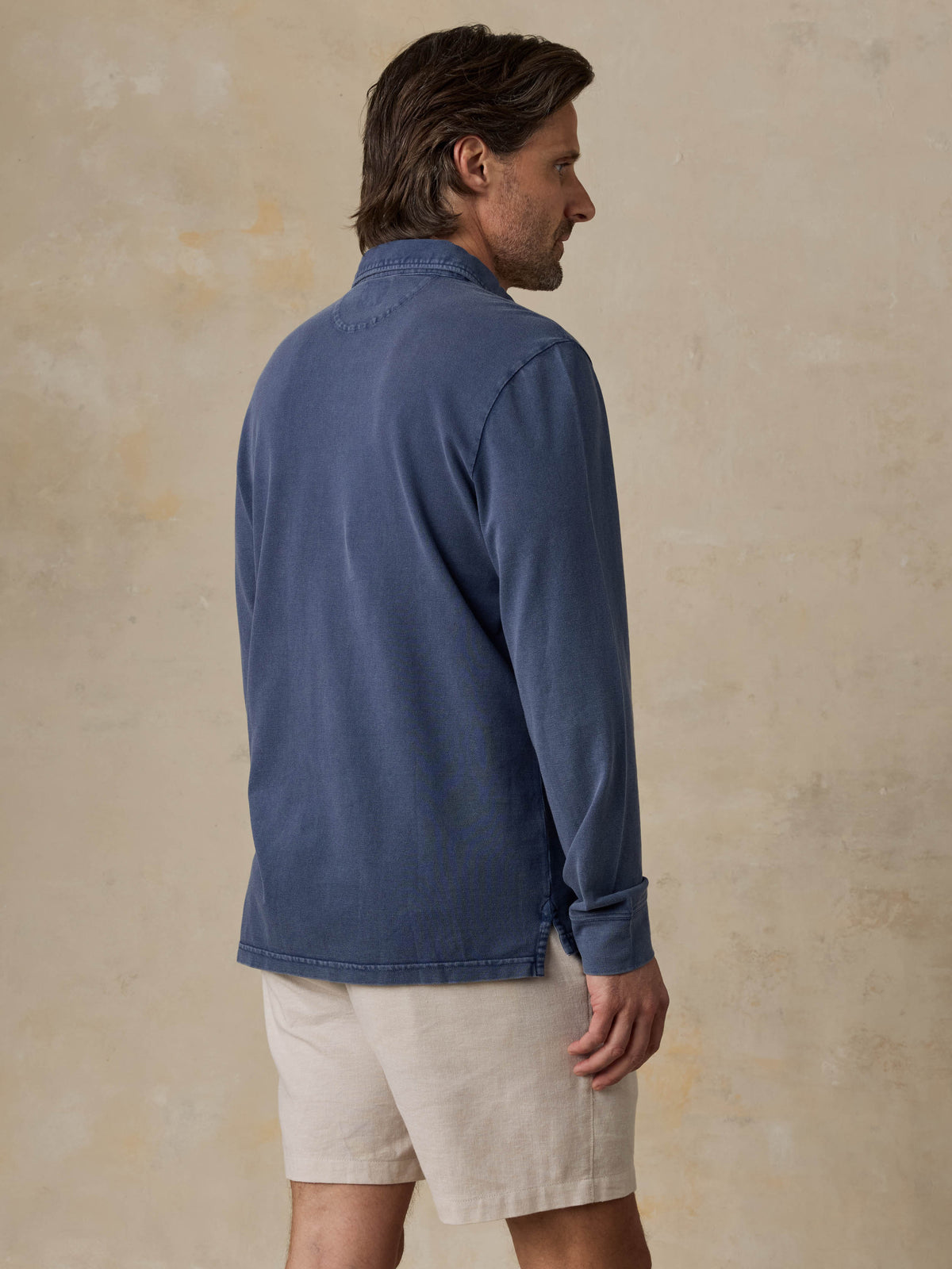 Man wearing a blue long-sleeve polo shirt and beige shorts against a beige background, image 4 of 6
