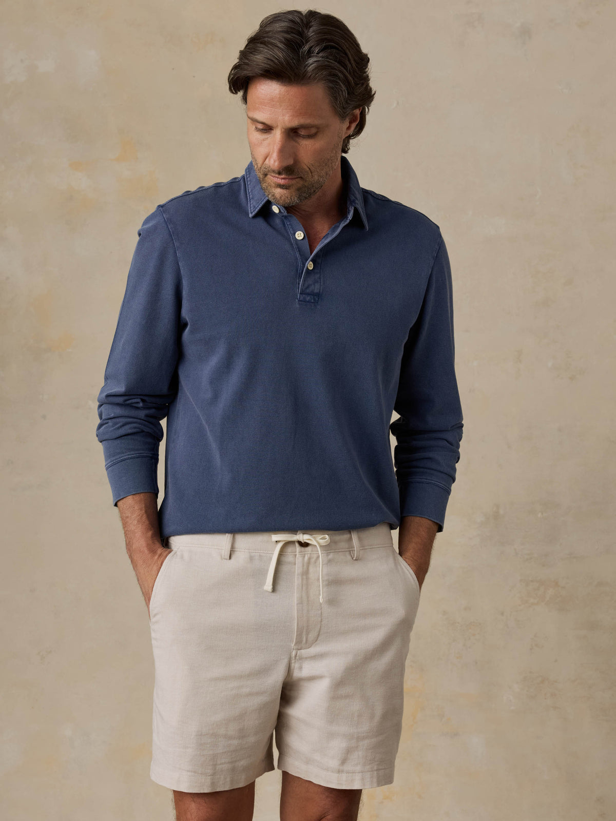 Man wearing a blue long-sleeve polo shirt and beige shorts against a beige background, image 1 of 6