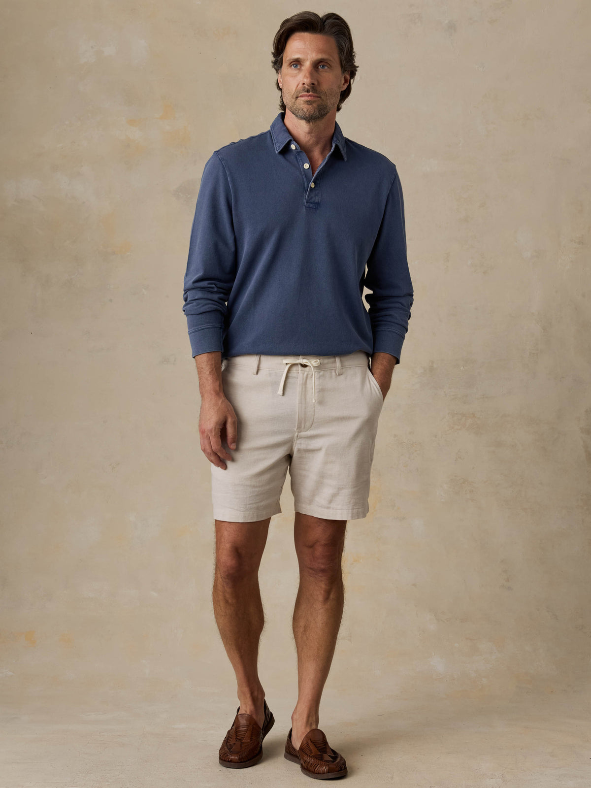 Man wearing a blue long-sleeve polo shirt and beige shorts against a beige background, image 6 of 6