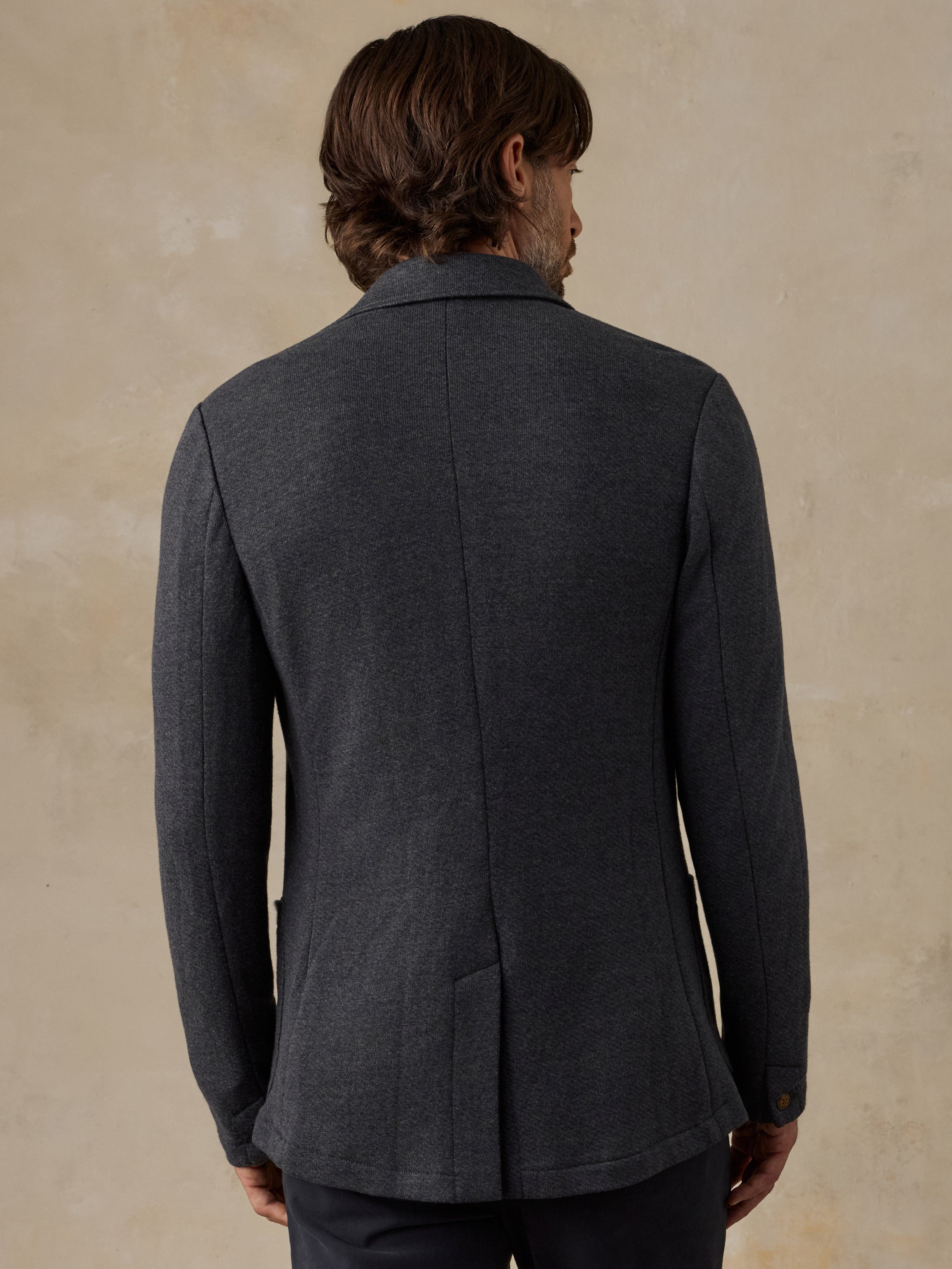 A man viewed from the back wearing a dark charcoal gray wool-blend blazer, set against a textured beige studio background.
