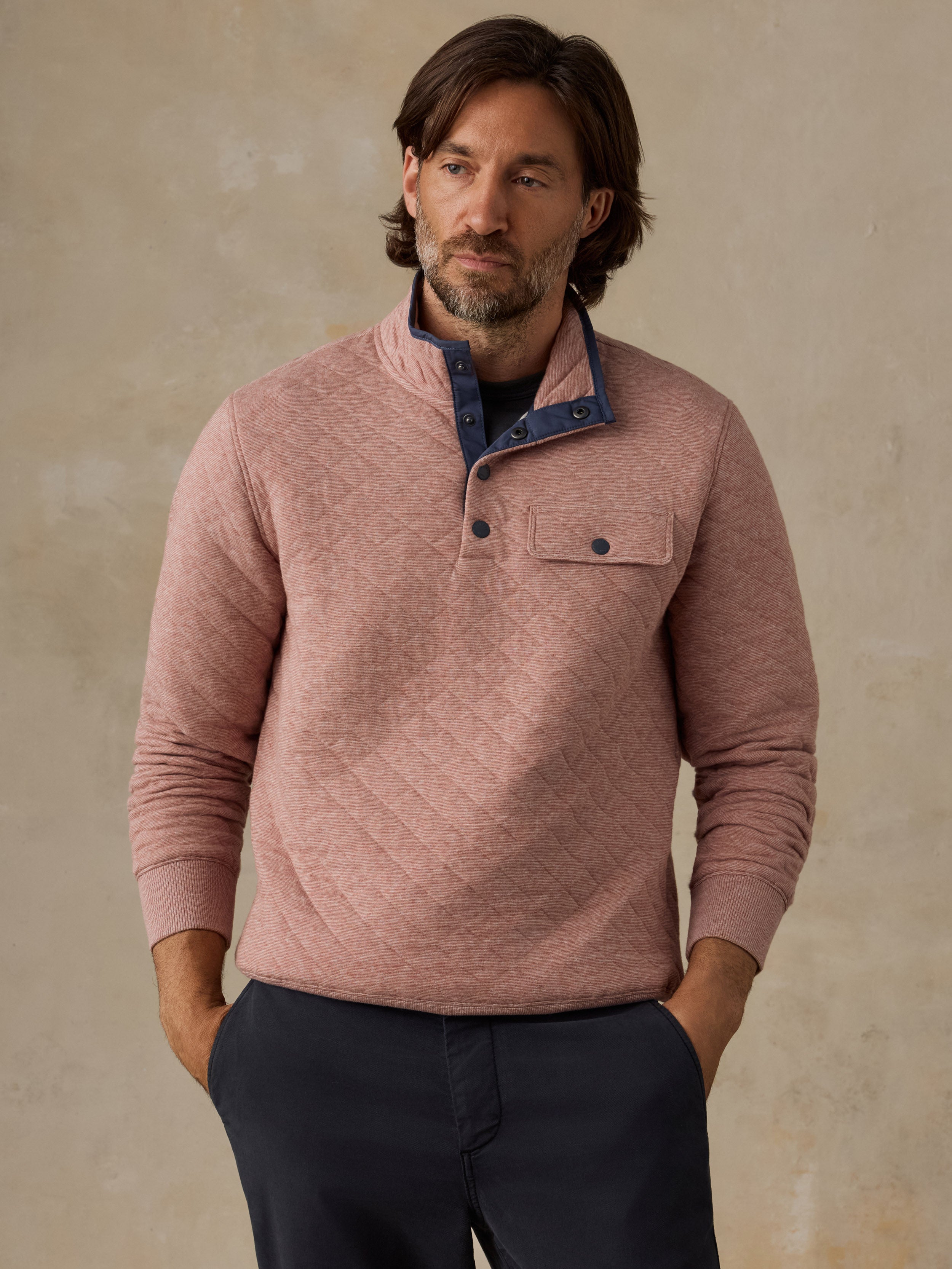 A man wearing a dusty rose quilted long-sleeve henley sweater with a dark blue inner collar and chest pocket, paired with dark navy pants, standing against a textured beige wall.