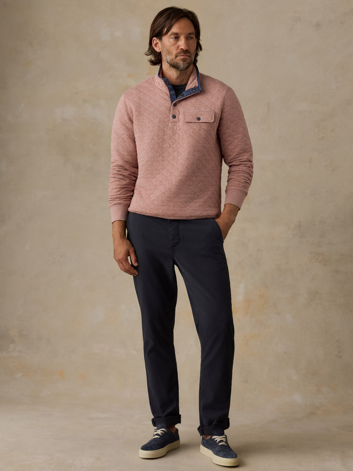 A man wearing a dusty pink long-sleeve quarter-placket sweater over a blue collared shirt, paired with navy pants and navy sneakers, standing against a textured beige studio backdrop., image 5 of 5