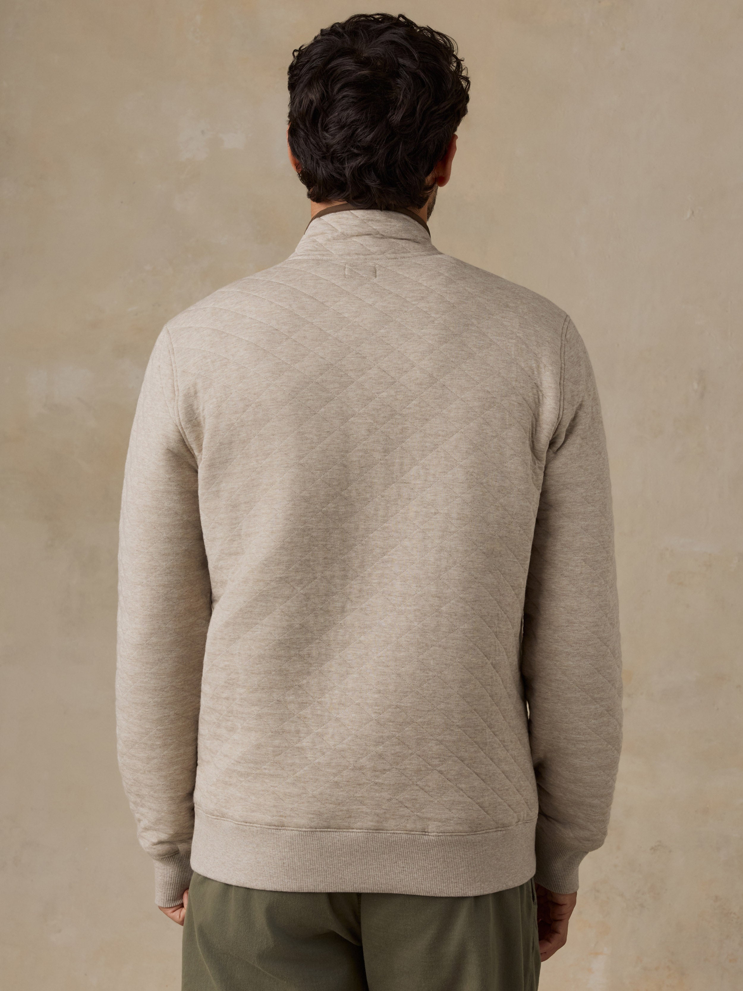 A man viewed from behind wearing a light beige knit fleece pullover and olive green pants, standing against a textured beige wall.