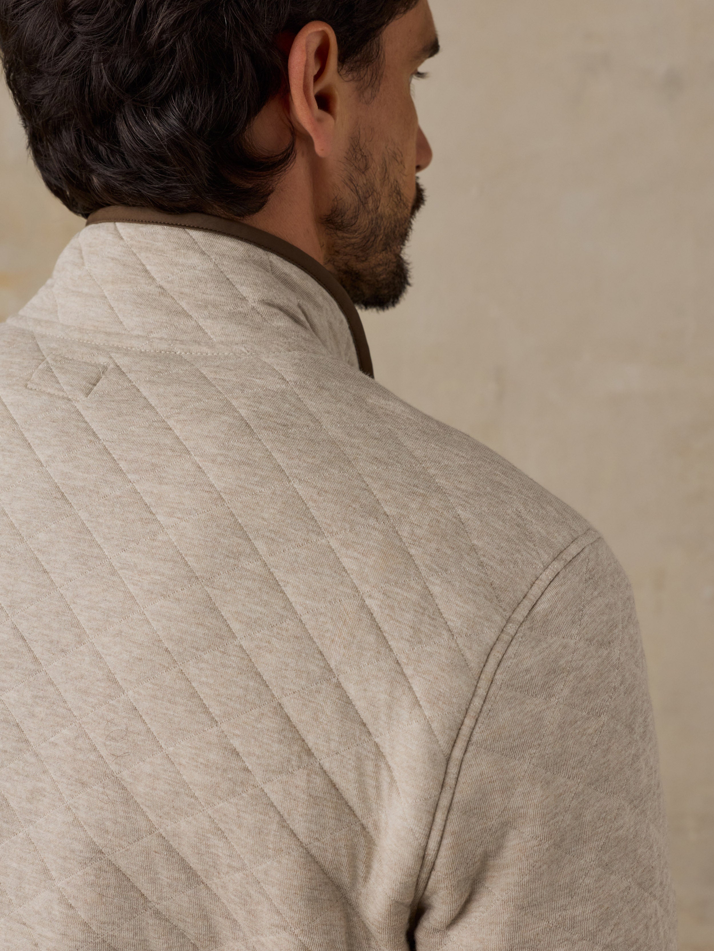 Close-up side profile of a man wearing a light beige quilted fleece pullover with a dark brown collar, set against a neutral textured beige wall.