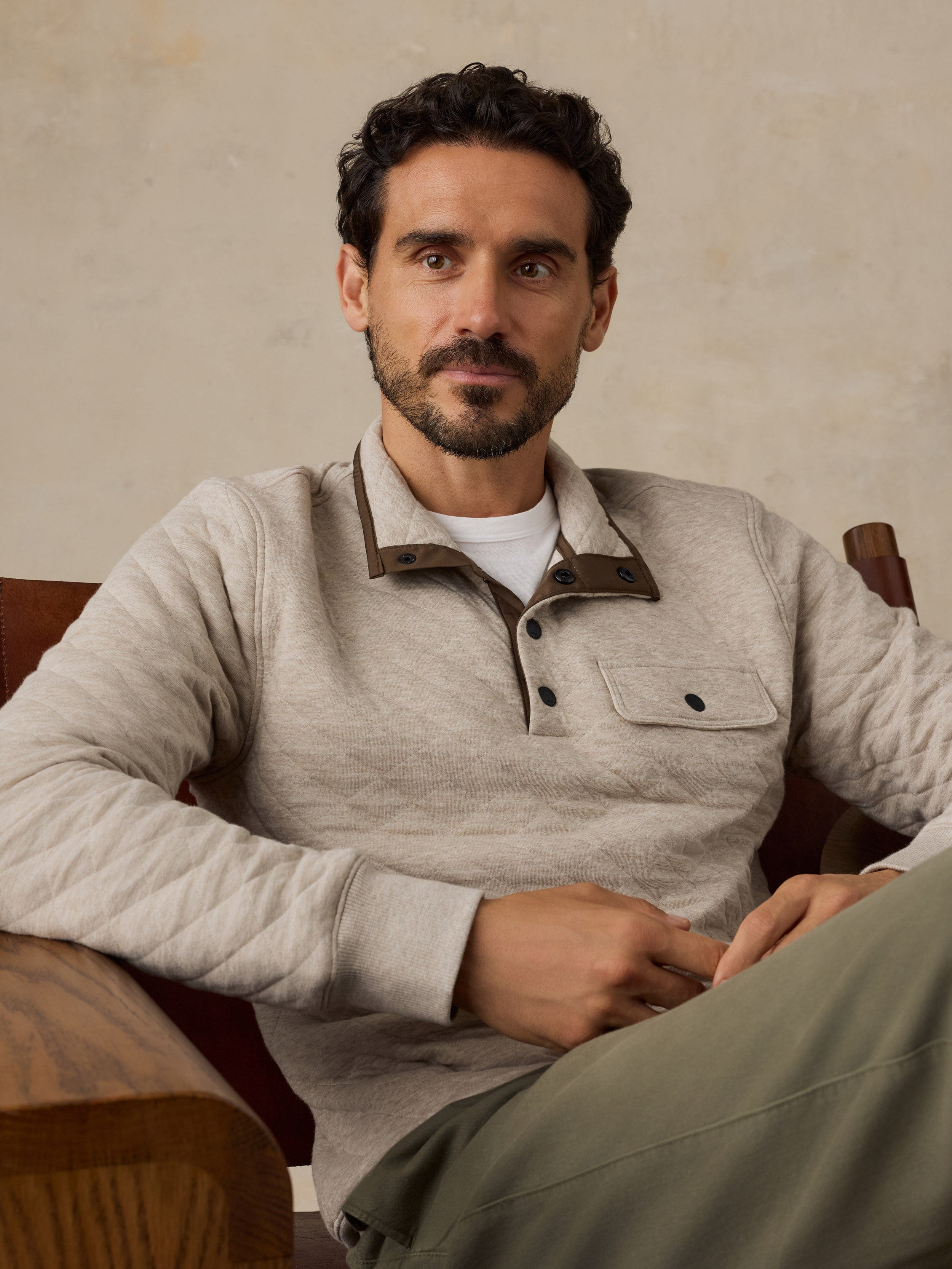A man wearing a light beige long-sleeve knit fleece pullover with brown contrast collar details, sitting in a wooden armchair against a textured beige wall.