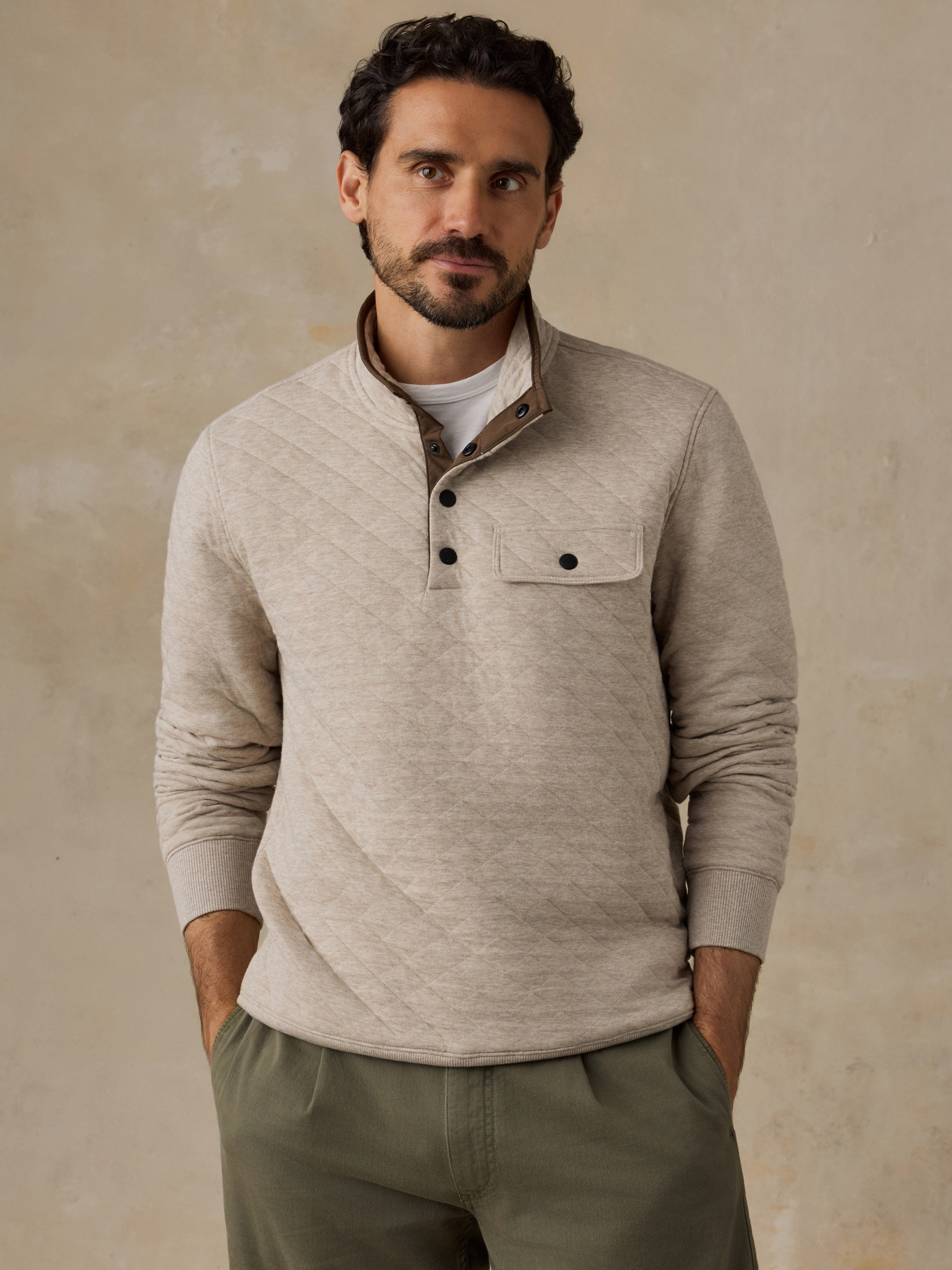 A man wearing a light beige long-sleeve fleece pullover with a buttoned collar and chest pocket, paired with olive green pants, standing against a textured beige background.