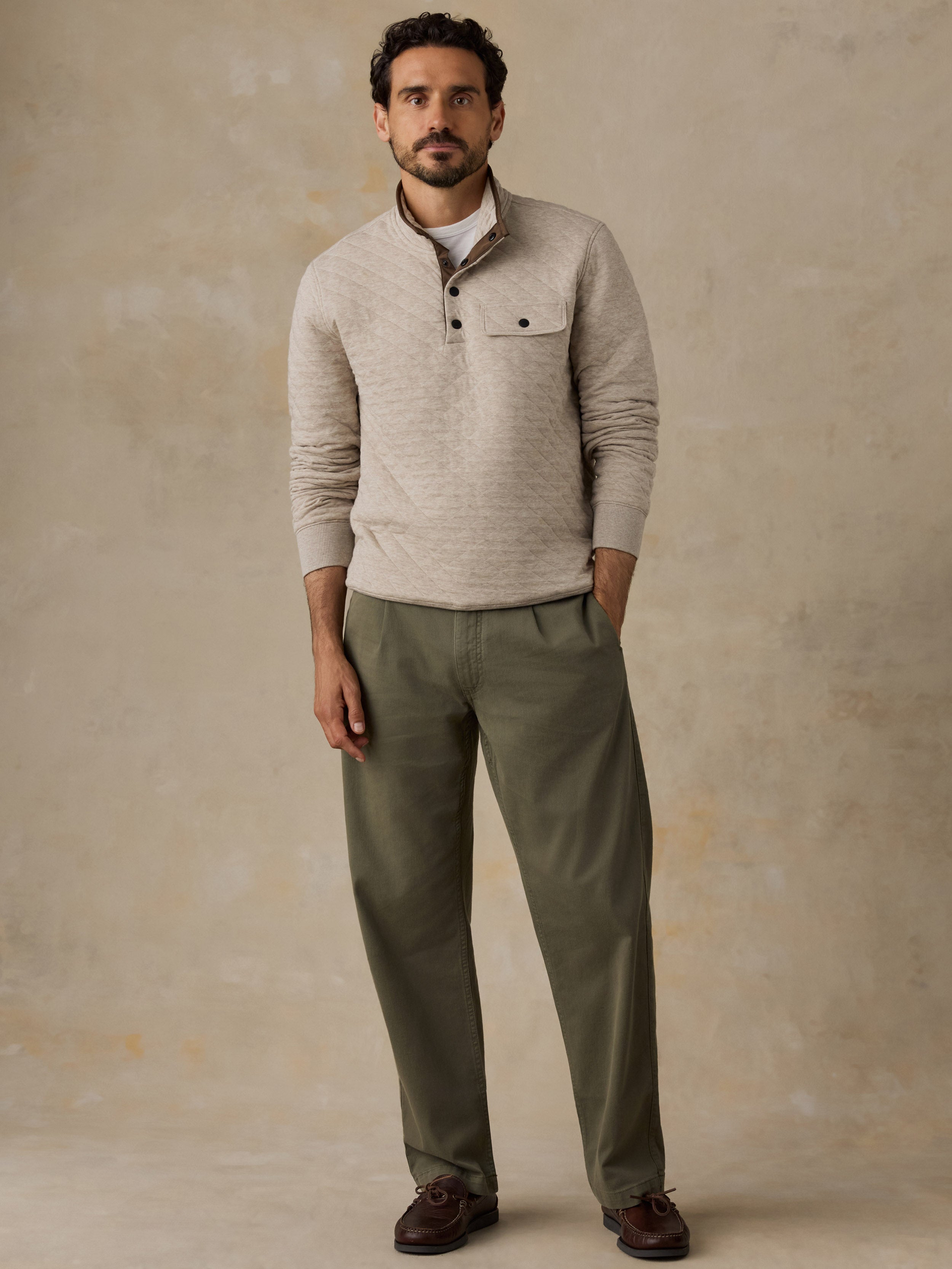 A man wearing a light beige textured long-sleeve fleece with a chest pocket and olive green trousers, standing against a textured beige studio backdrop.