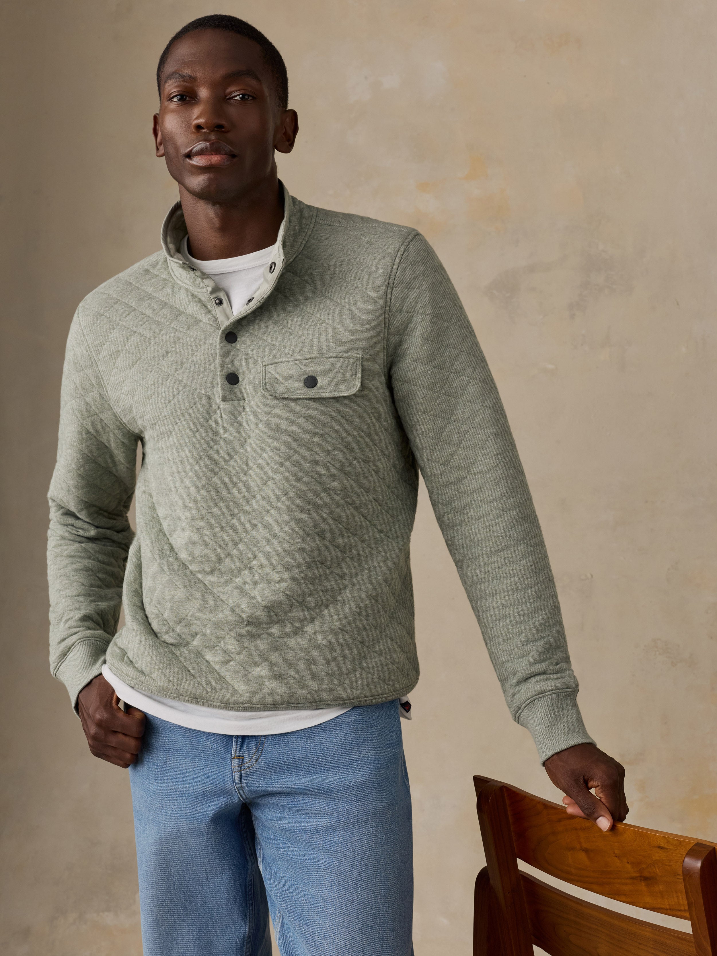 A man wearing a sage green quilted long-sleeve pullover over a white t-shirt and blue jeans, standing next to a wooden chair against a textured beige wall.