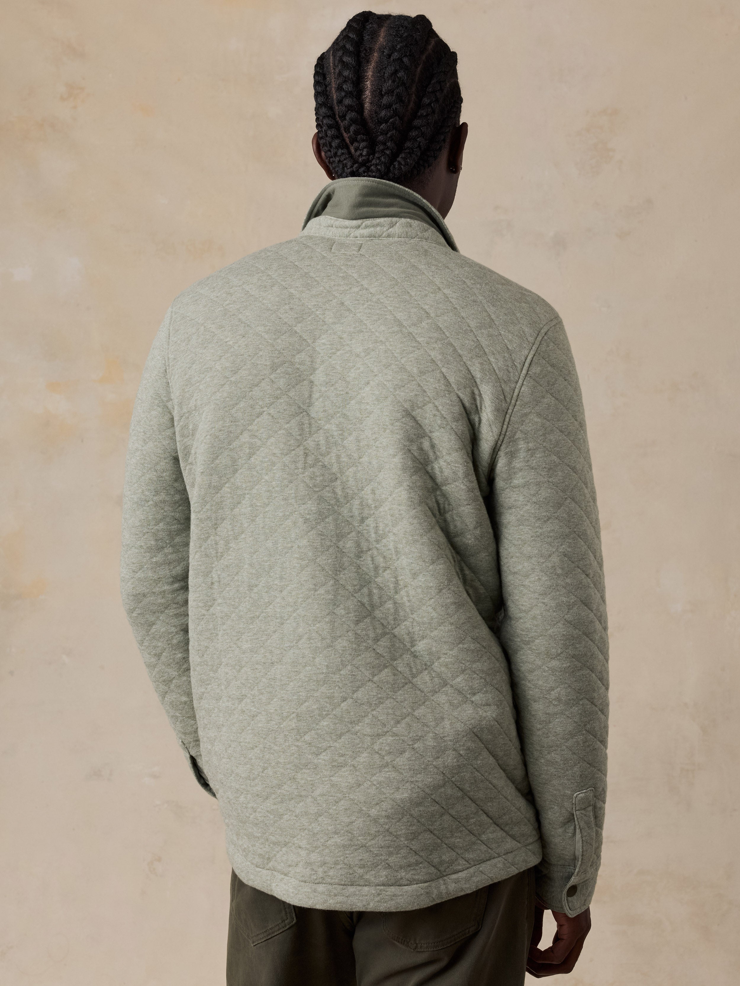 A man viewed from behind wearing a light sage green quilted fleece jacket and dark olive pants, standing against a textured beige wall.