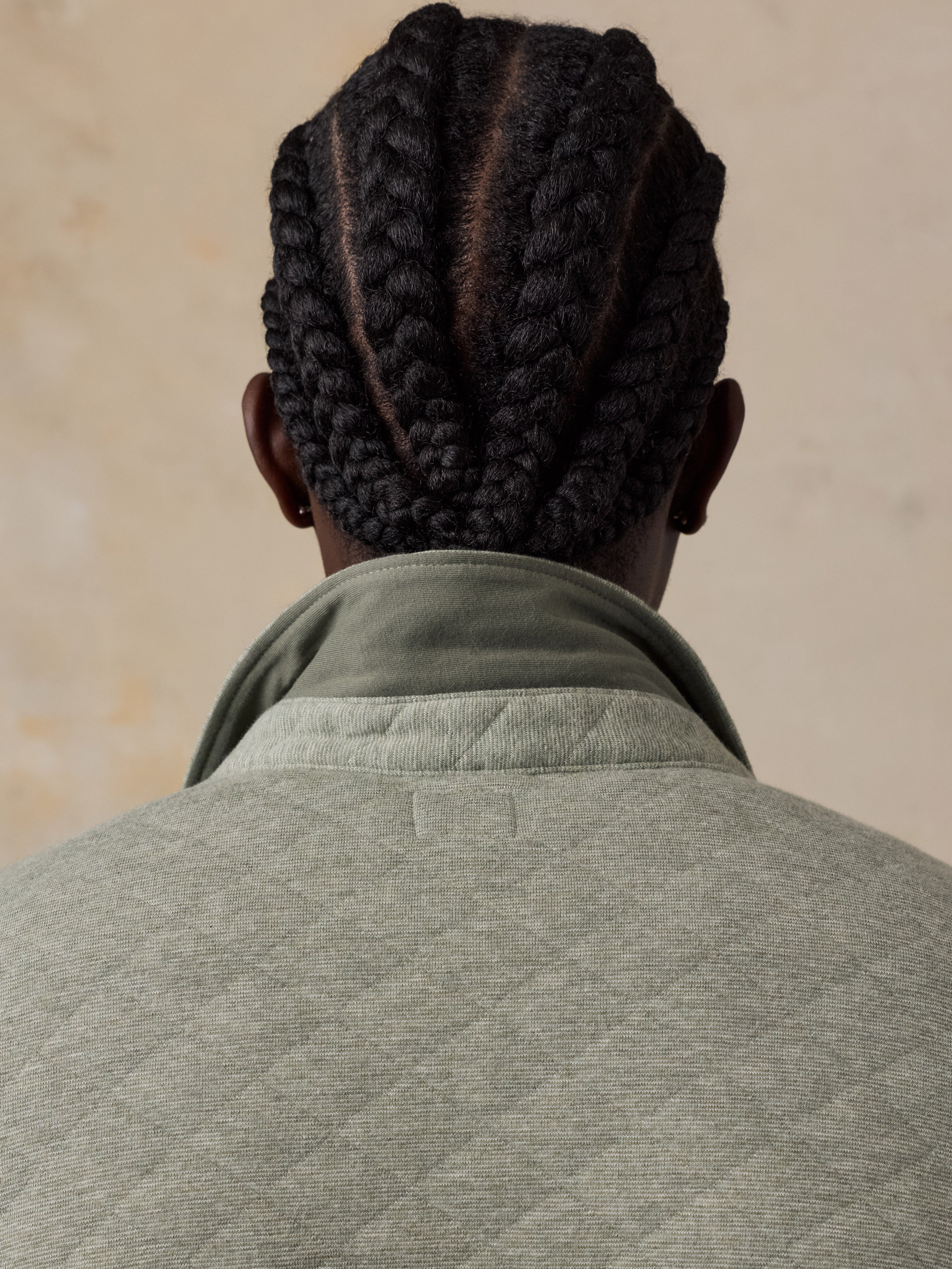 Back view of a man with dark braided hair, wearing a light gray quilted jacket over an olive green shirt, set against a neutral beige background.