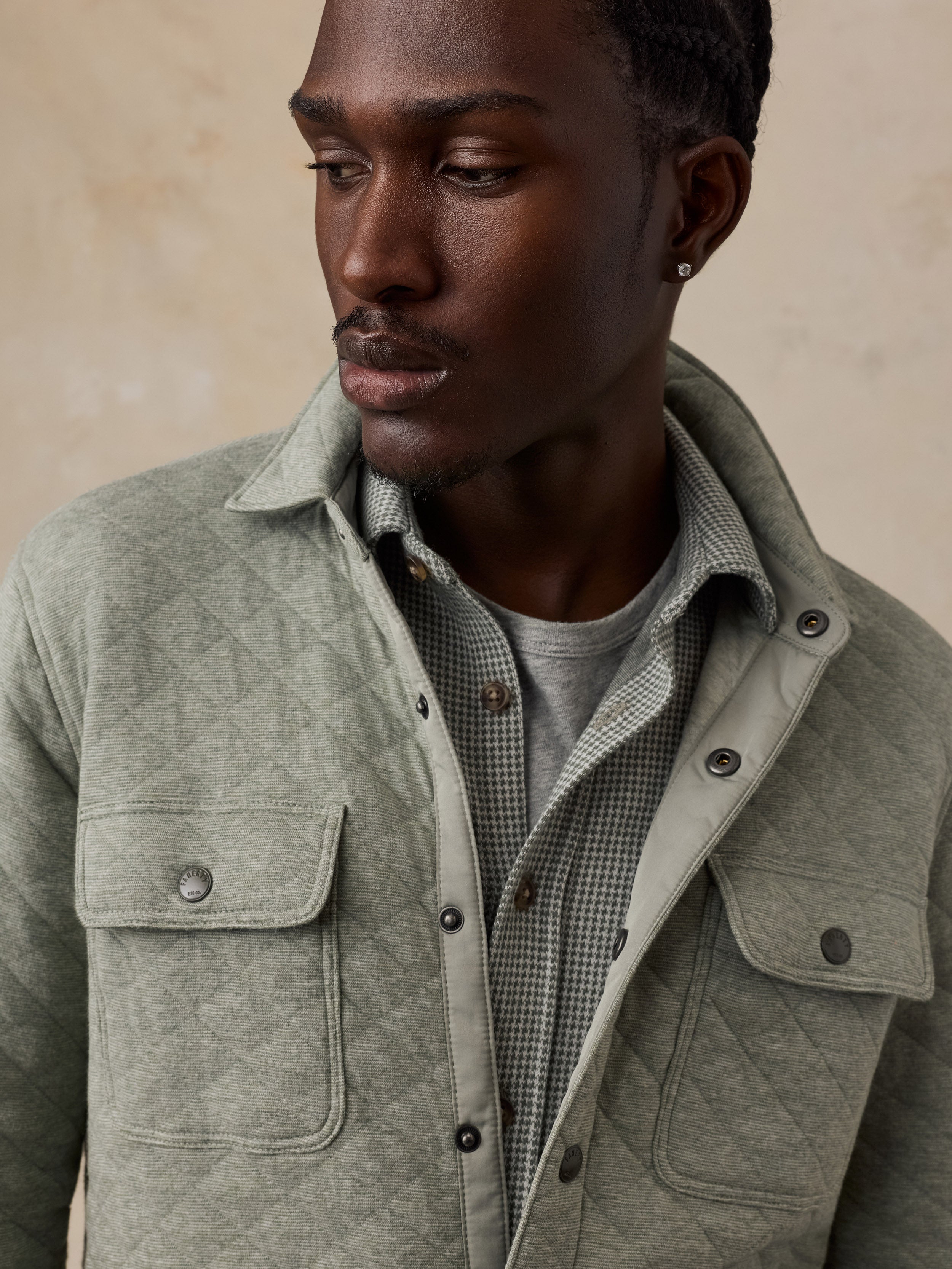 A close-up of a man wearing a sage green quilted overshirt over a patterned button-down shirt and a gray t-shirt, set against a warm beige textured background.