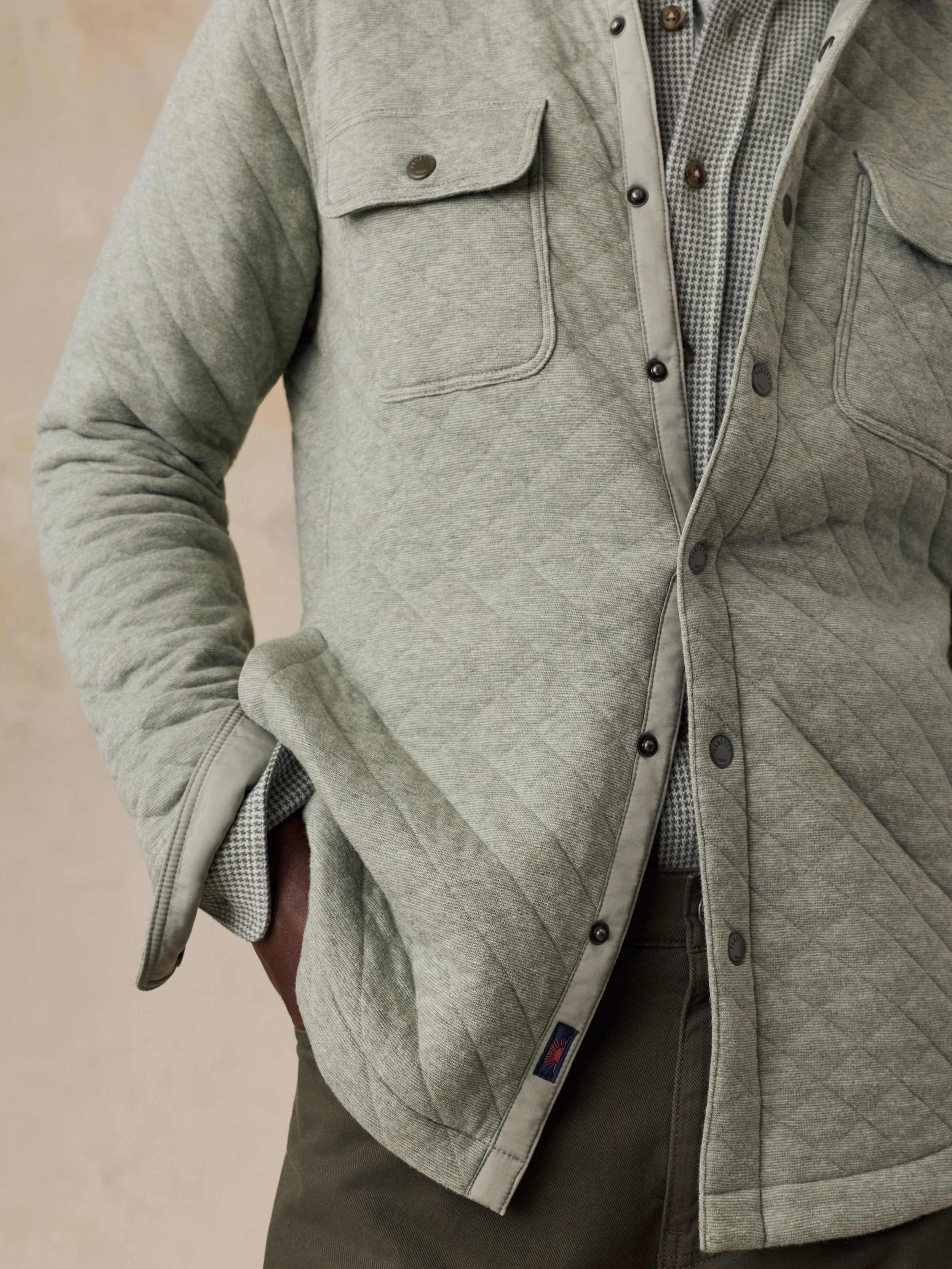 A close-up of a man wearing a light gray quilted overshirt jacket over a checkered shirt and dark trousers, with one hand in a pocket.