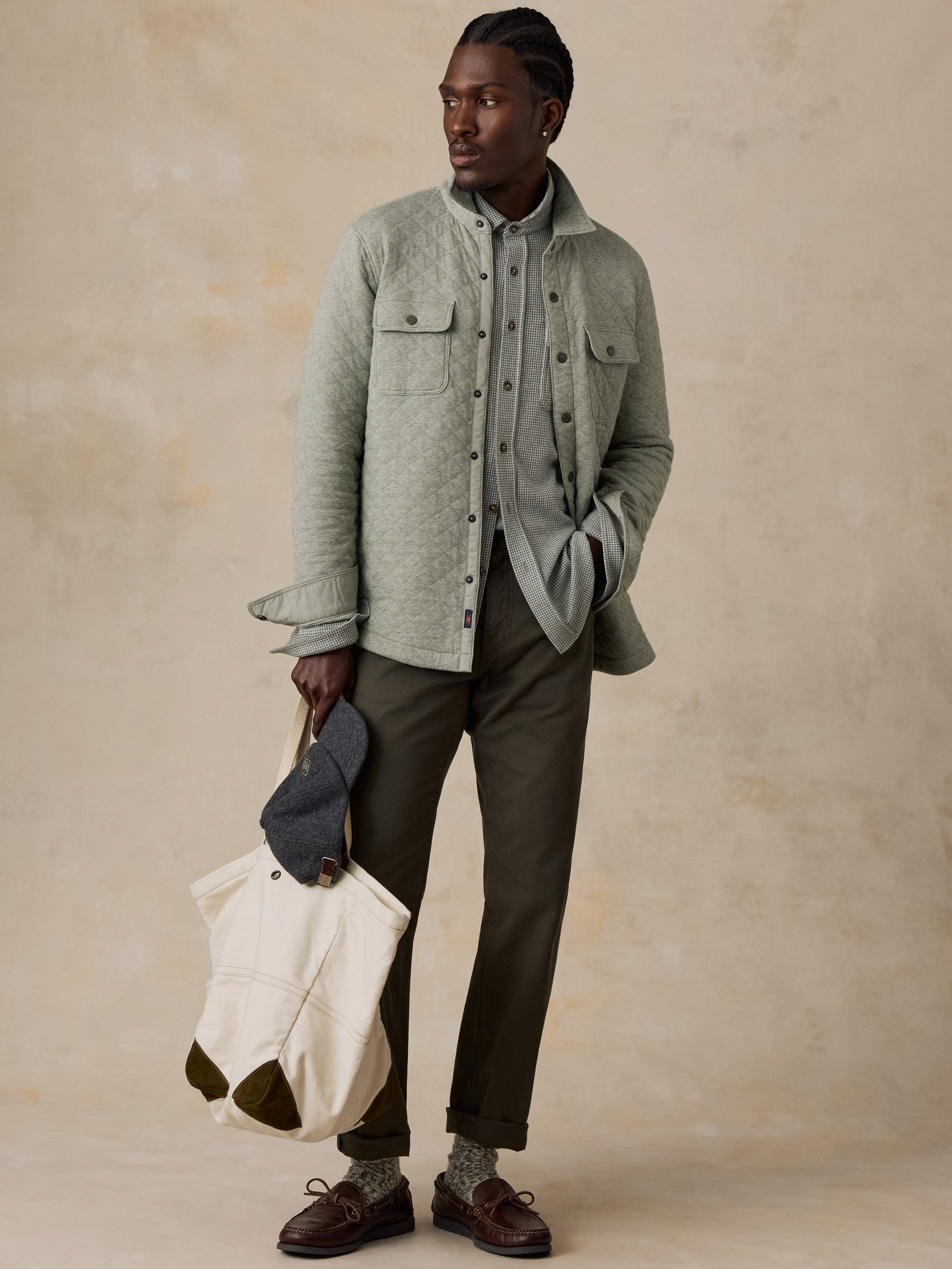 A man stands against a textured beige studio backdrop wearing a light sage green overshirt over a gray button-up shirt, dark olive trousers, brown shoes, and holding a cream tote bag with a gray cap.