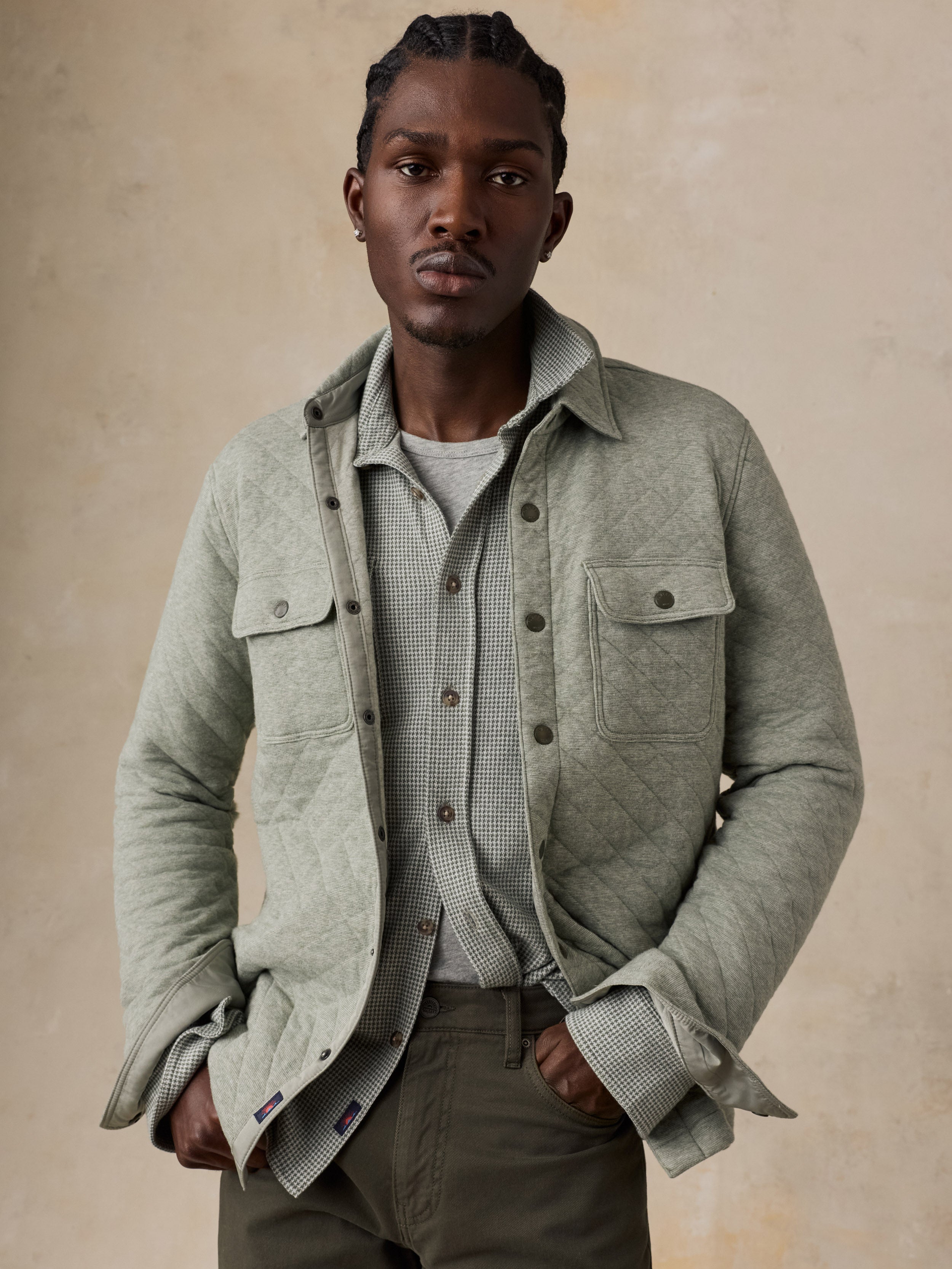 A man wearing a light sage green quilted overshirt jacket over a gray patterned button-up shirt and olive pants, standing against a textured beige background.