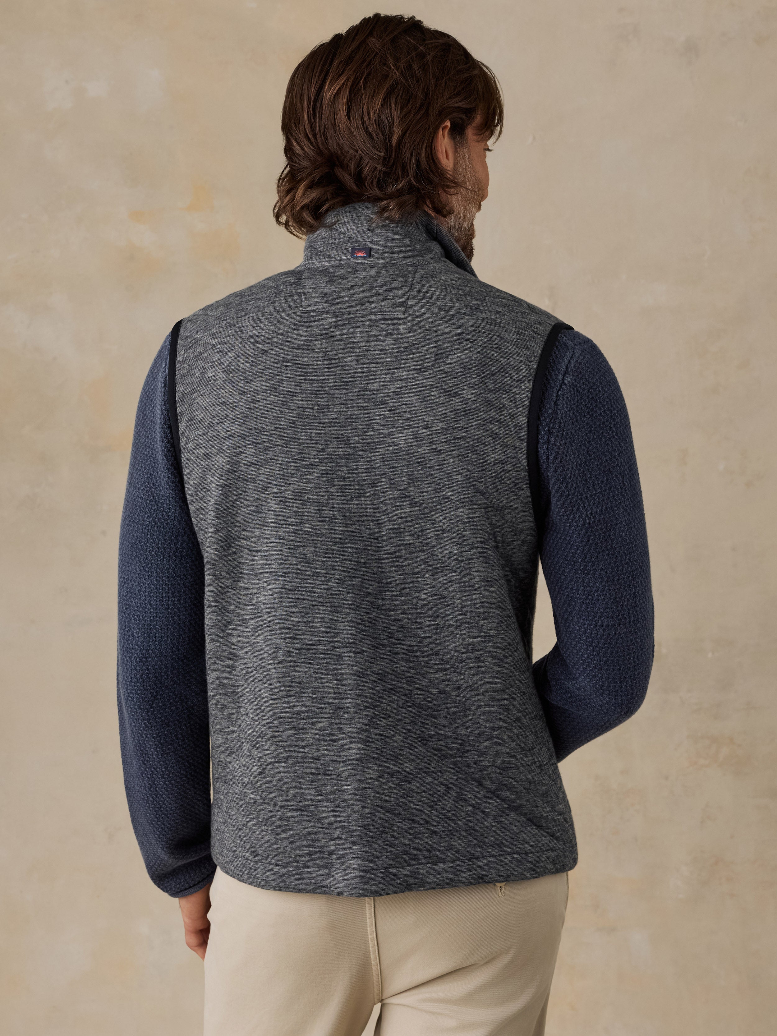 A man shown from the back wearing a heather gray ribbed quarter-zip vest over a navy blue long-sleeve knit sweater, paired with beige pants, against a textured beige background.