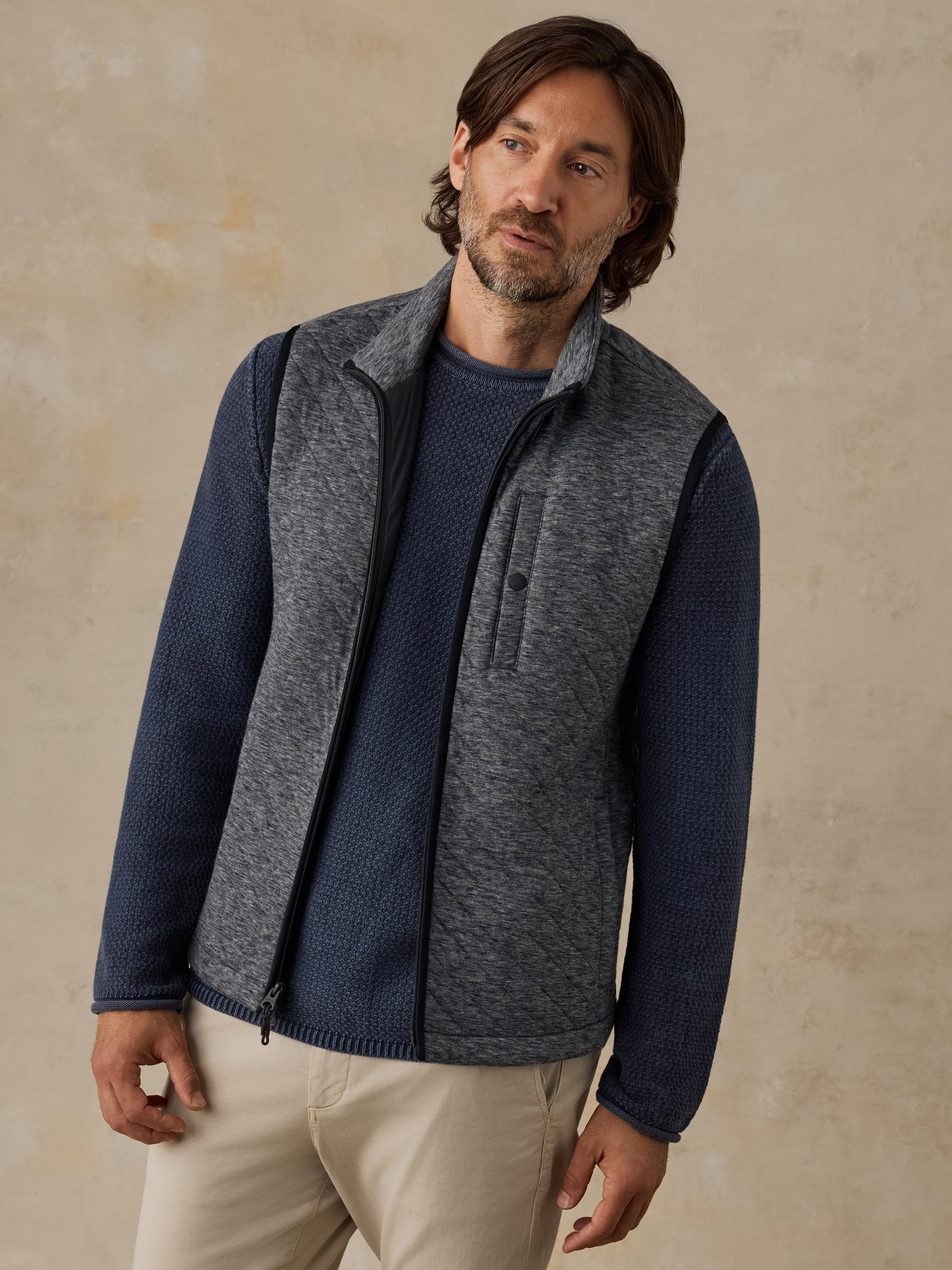 A man wearing a gray heather zip-front vest over a navy blue knit sweater and beige pants, standing against a textured beige studio background.
