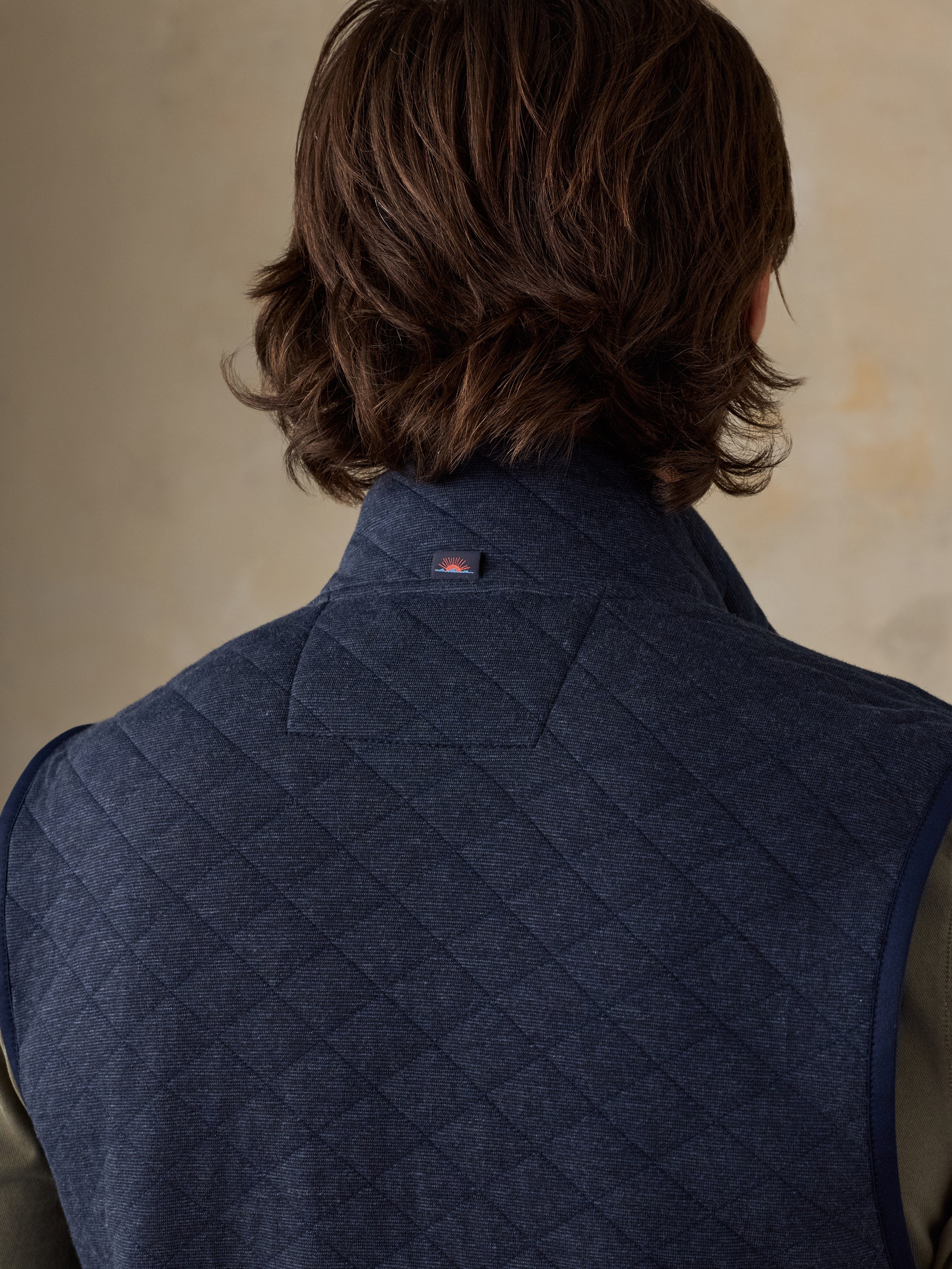 Back view of a man wearing a navy blue quilted waistcoat over a dark shirt against a beige background.