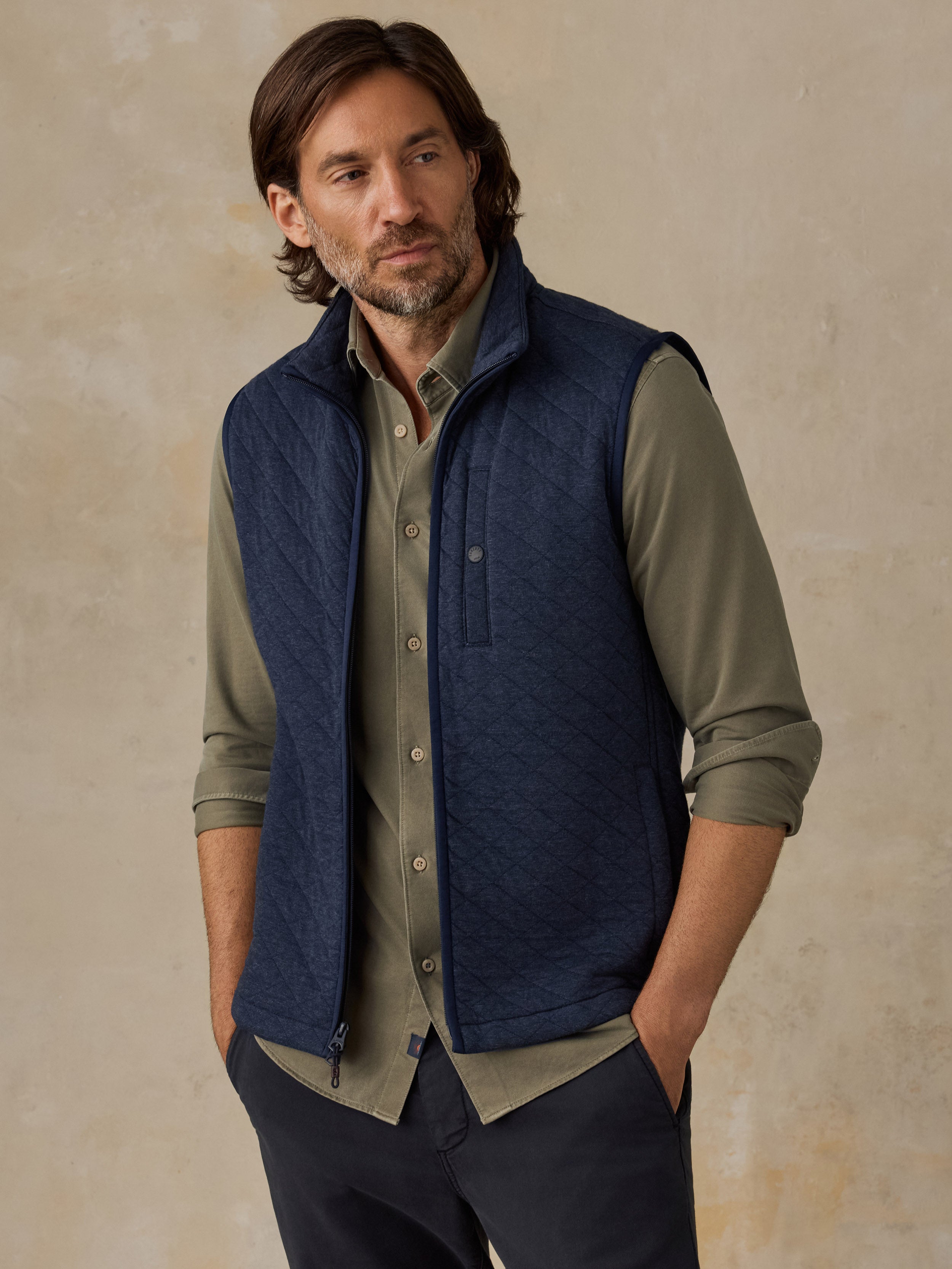 A man wearing a navy blue quilted vest over an olive green button-down shirt, paired with dark trousers, standing against a textured beige studio backdrop.