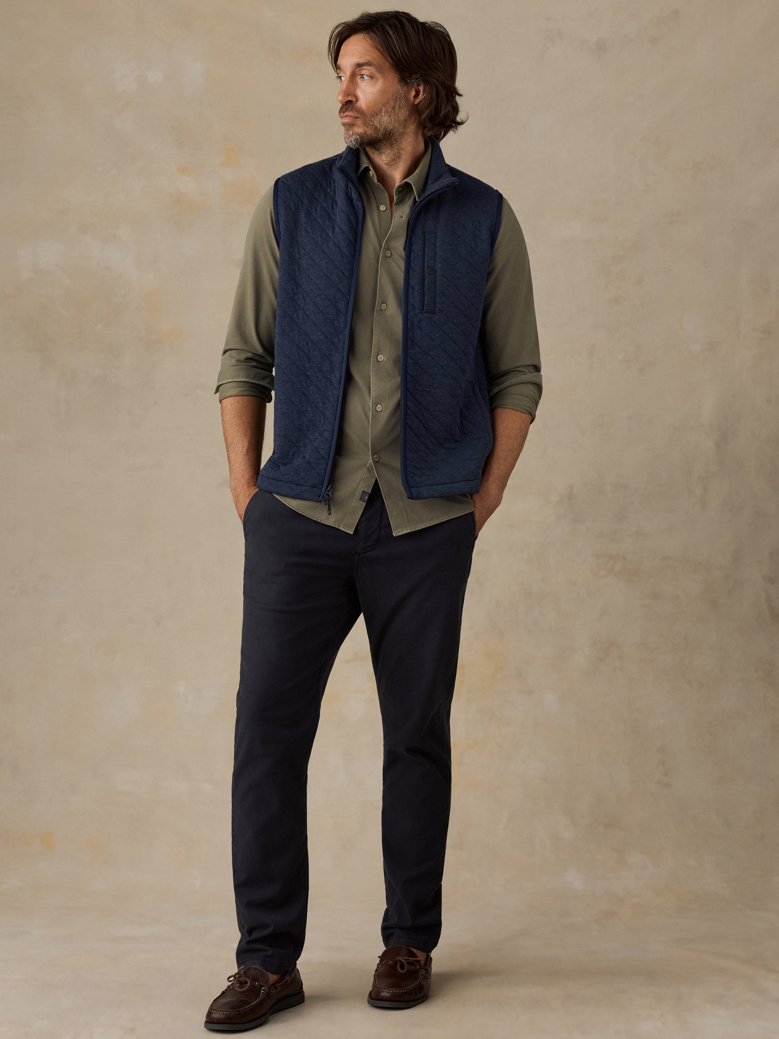 A man wearing a navy blue quilted vest over an olive green button-down shirt, paired with dark navy pants and brown shoes, standing against a textured beige studio backdrop.