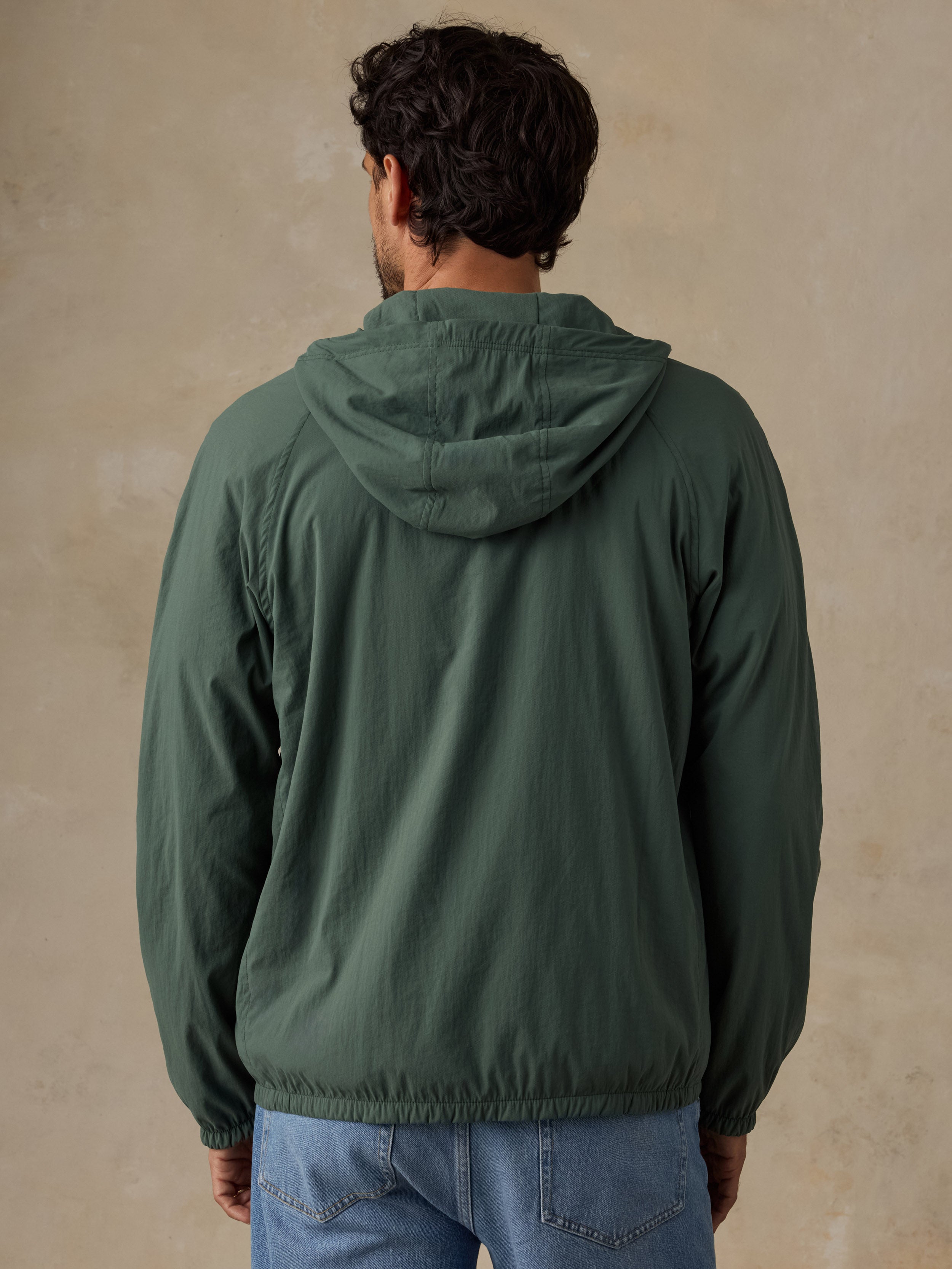 A man viewed from behind wearing a dark green hooded windbreaker jacket and blue jeans, standing against a textured beige wall.