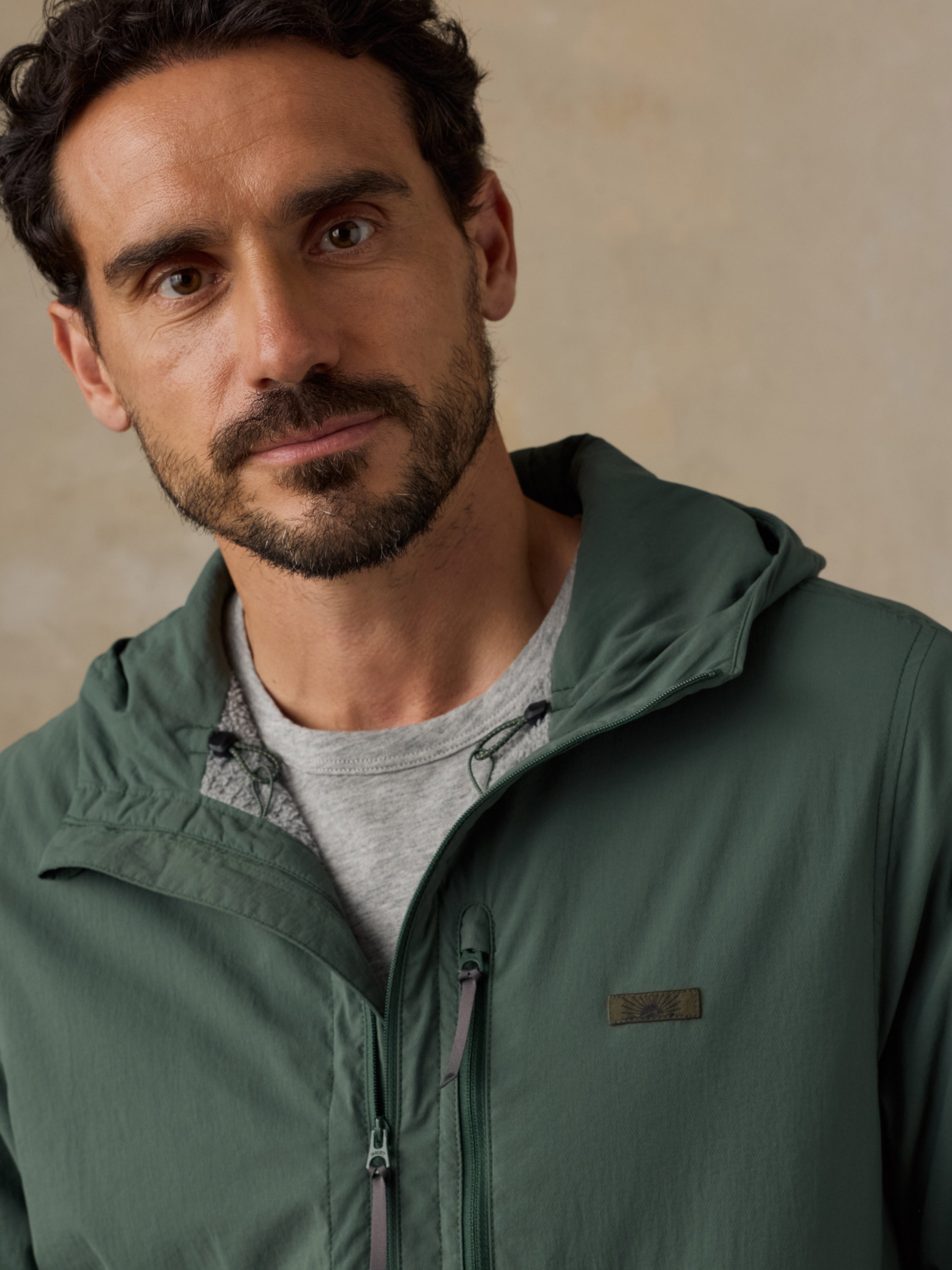 A close-up shot of a man wearing a sage green hooded lightweight jacket over a light gray t-shirt, set against a neutral textured beige background.