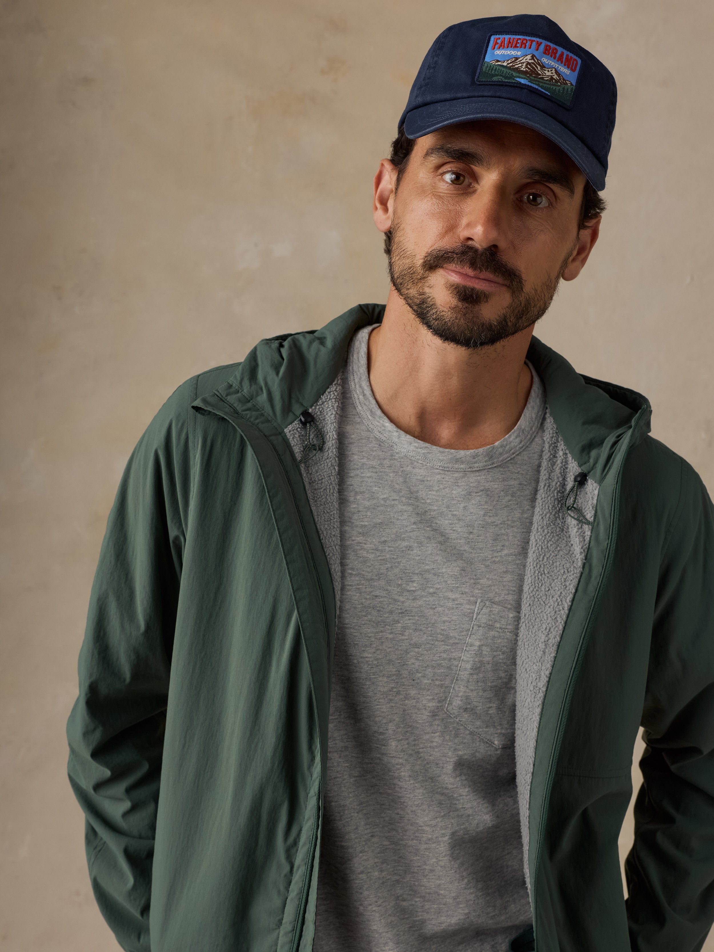Man wearing a blue baseball hat, green jacket, and a grey pocket t-shirt against a beige background