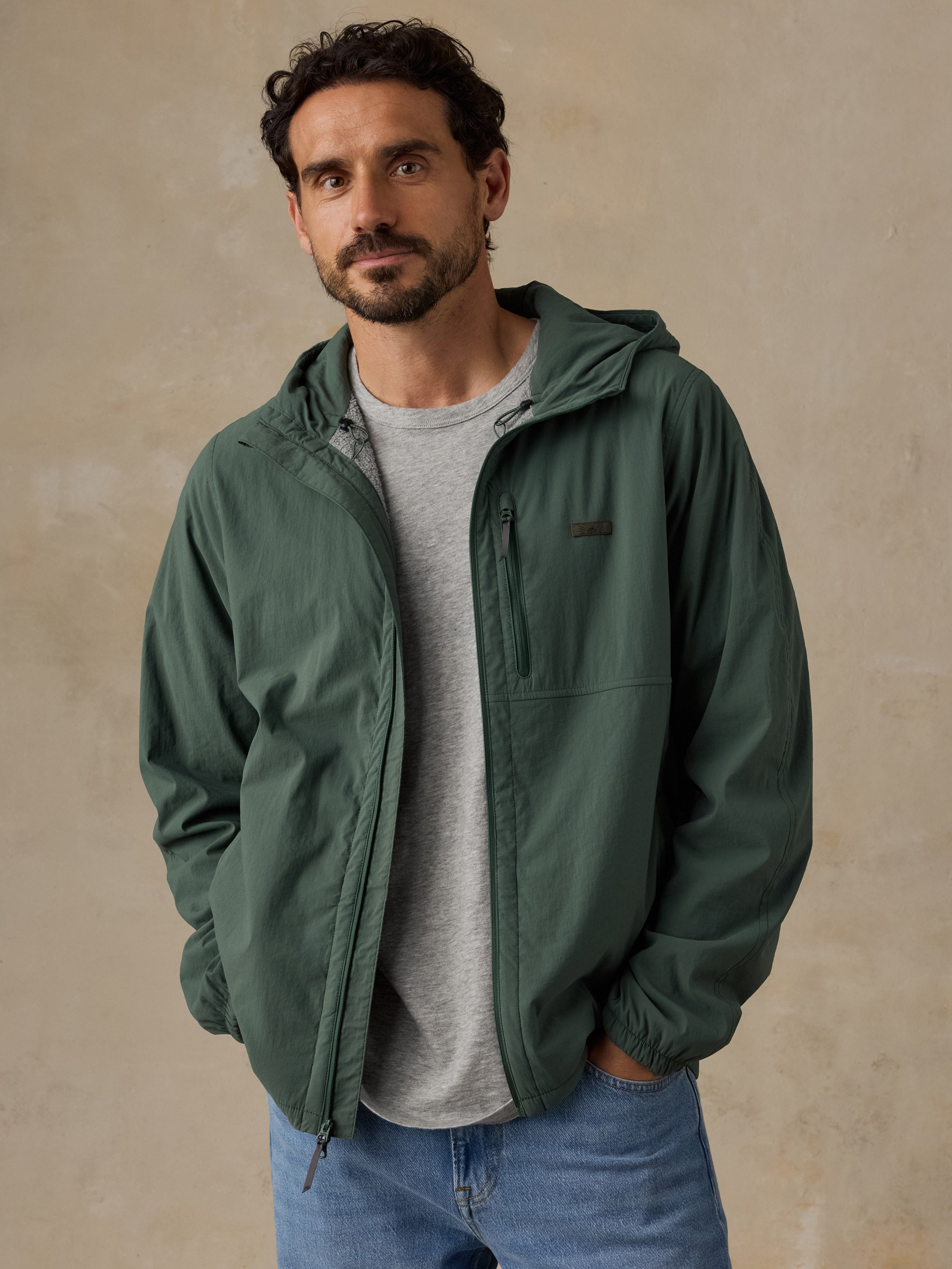 A man wearing a dark green hooded jacket over a light gray t-shirt and blue jeans, standing against a textured beige wall.