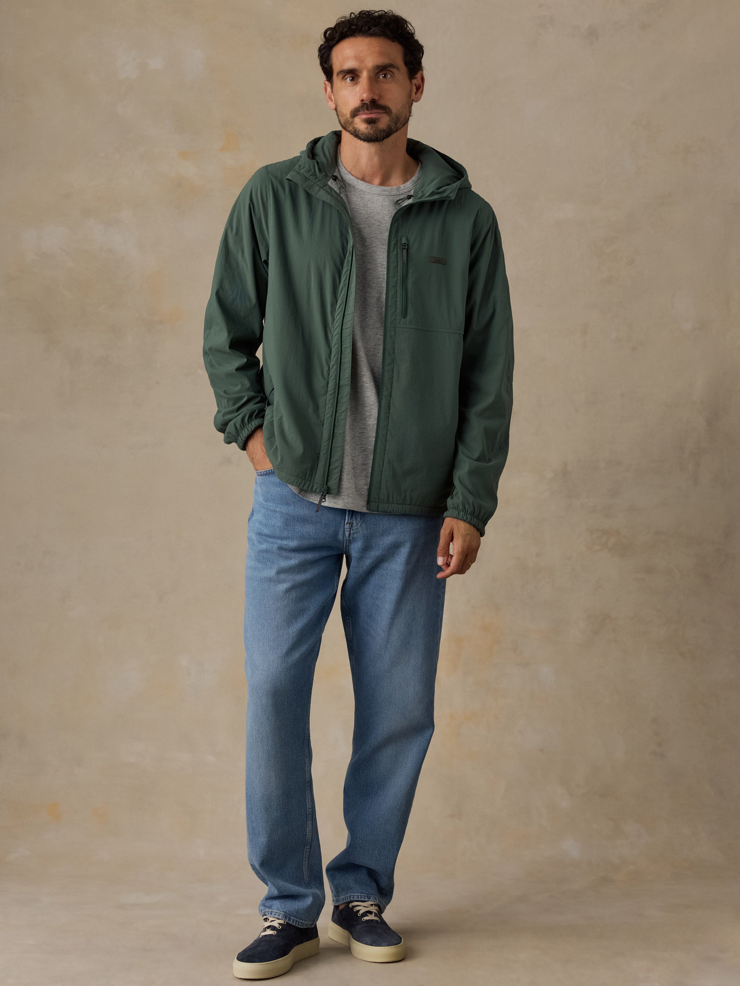Man wearing a forest green jacket over a grey pocket t-shirt and blue jeans against a beige background