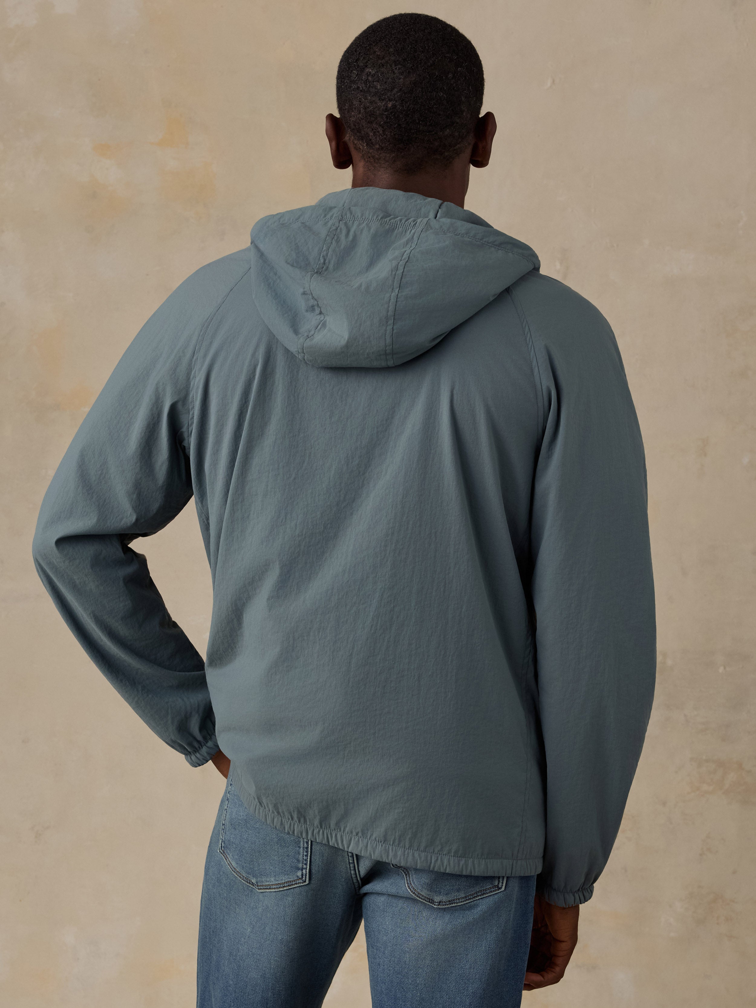 A man viewed from behind wearing a muted blue-gray hooded jacket and blue jeans, standing against a textured beige wall.