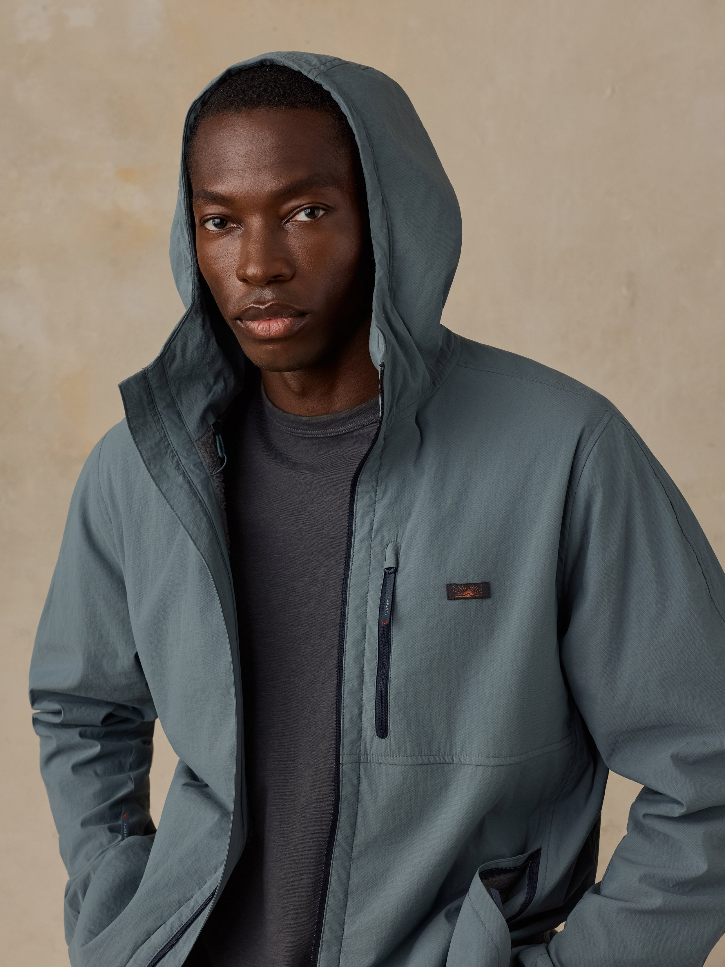 A man wearing a slate blue-gray hooded jacket over a dark gray t-shirt, standing against a textured beige background.