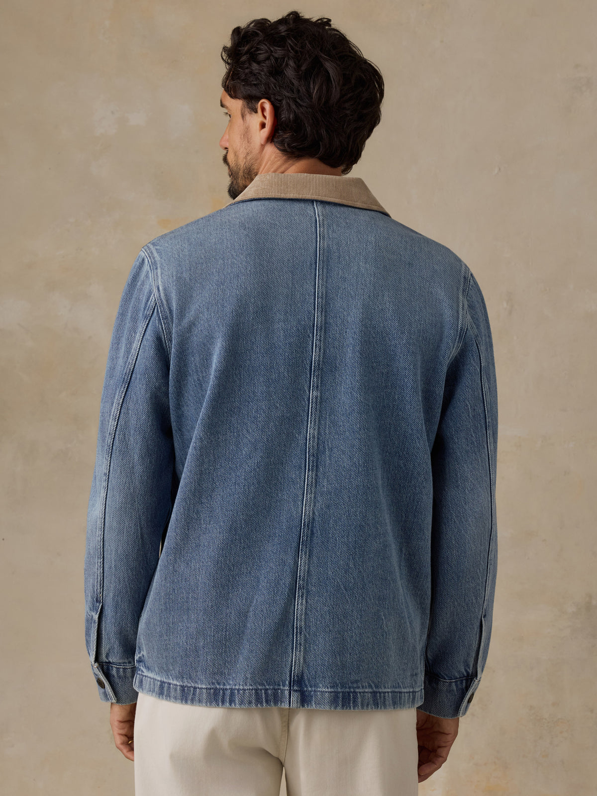 Man wearing a blue denim jacket with tan corduroy collar and white chino pants against a beige background, image 4 of 6