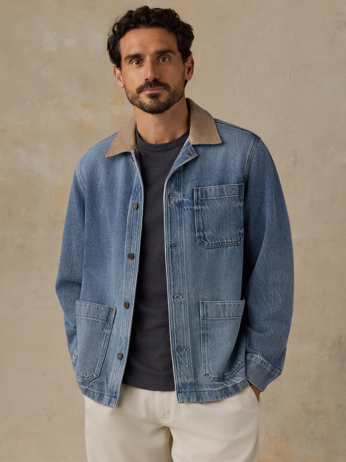Man wearing a blue denim jacket with tan corduroy collar, blue t-shirt, and white chino pants against a beige background, image 1 of 6