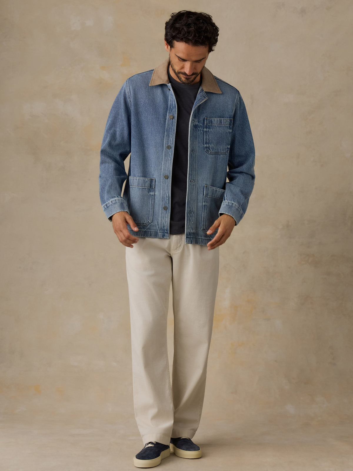 Man wearing a blue denim jacket with tan corduroy collar, blue t-shirt, and white chino pants against a beige background, image 6 of 6