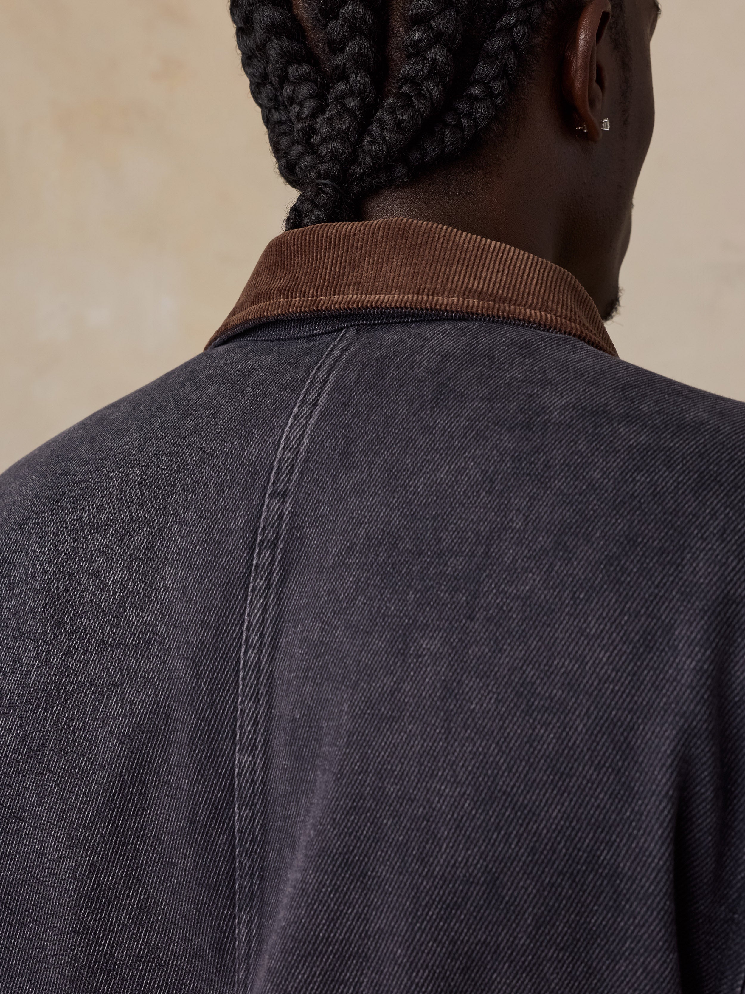 Close-up back view of a man wearing a dark charcoal gray textured jacket with a brown corduroy collar, against a warm beige background.