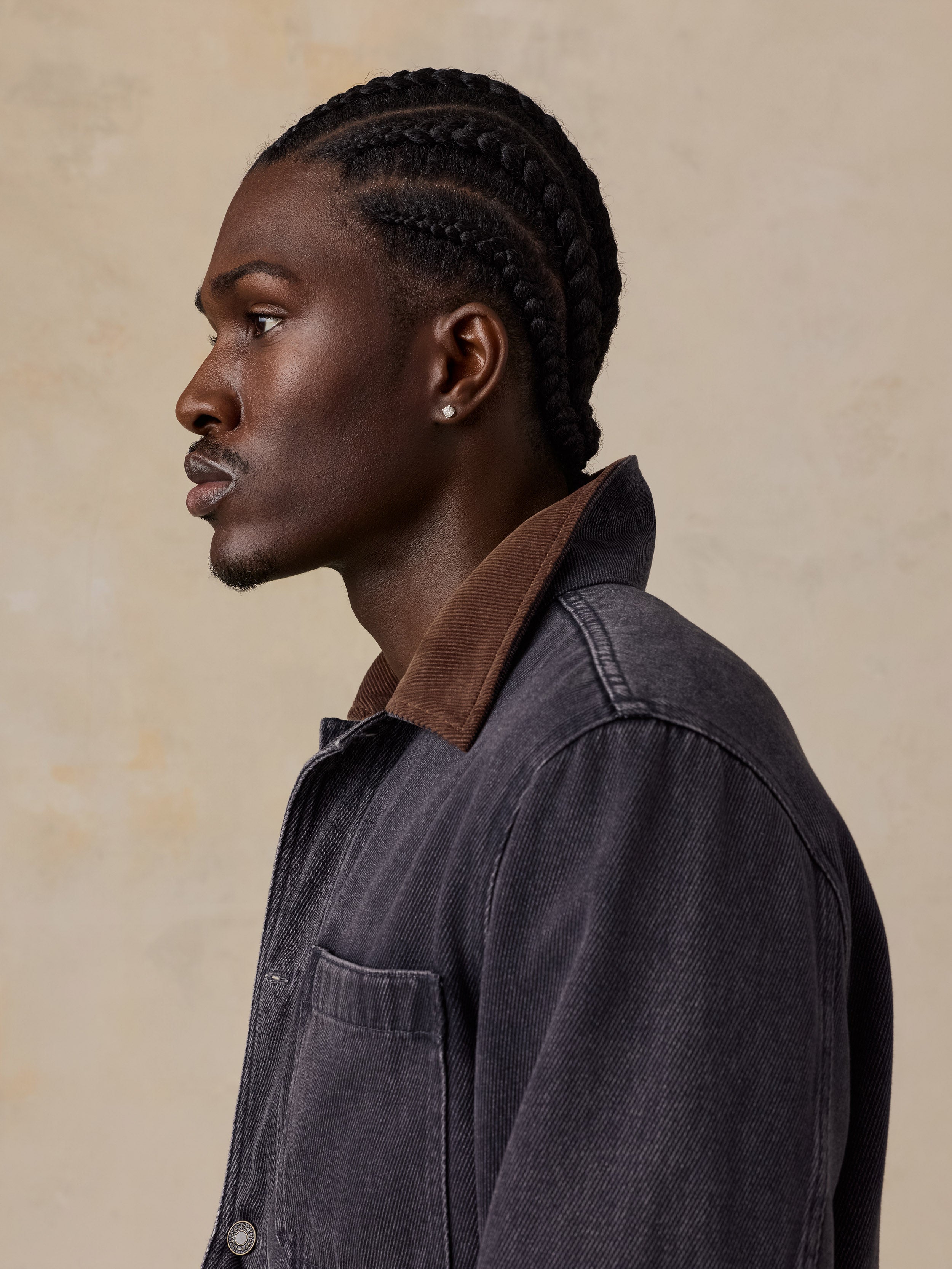A man in profile wearing a dark charcoal gray corduroy jacket with a brown corduroy collar, set against a textured beige background.