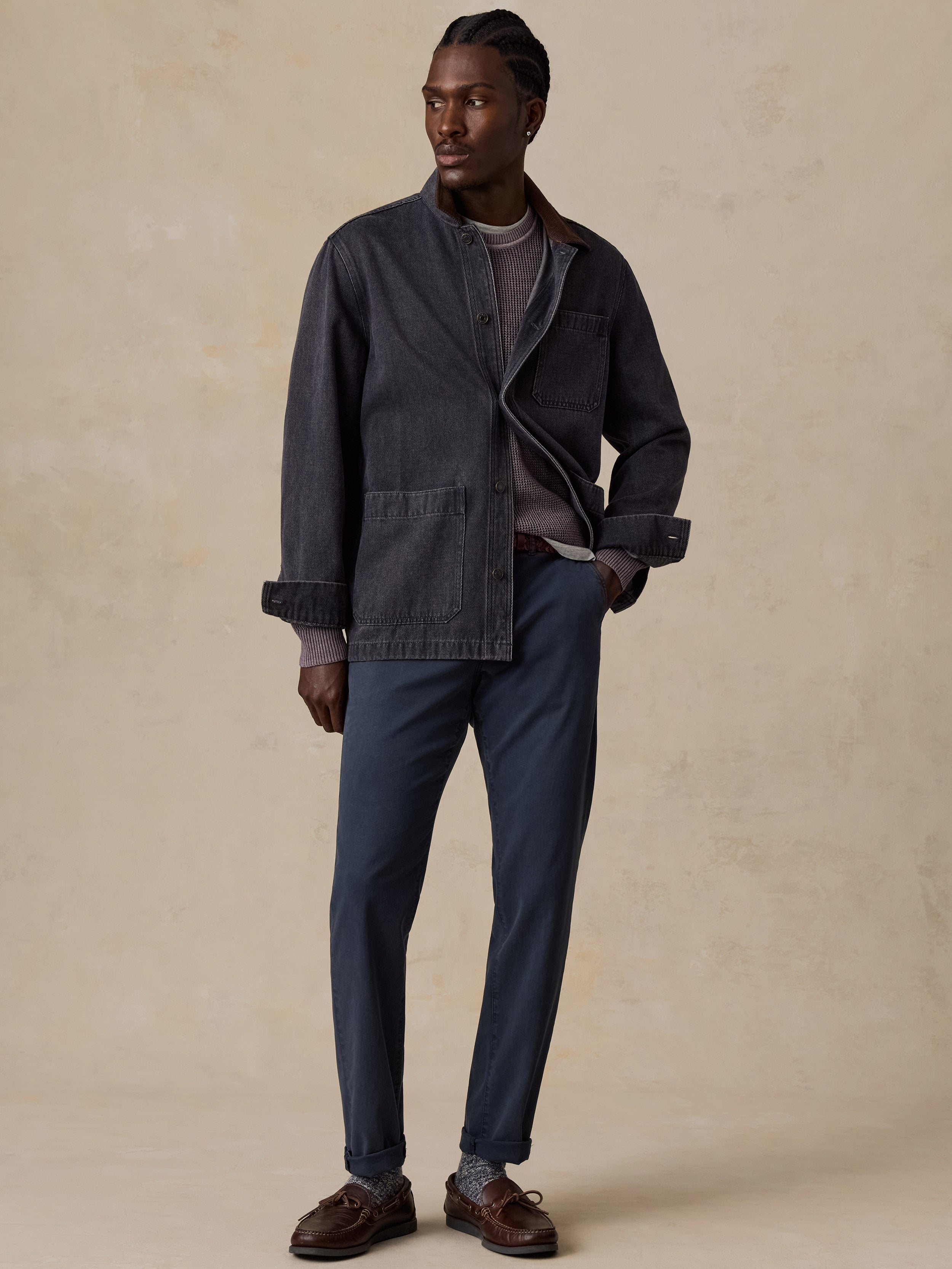 A man modeling a dark charcoal gray overshirt jacket, navy blue trousers, a gray t-shirt, and brown leather shoes against a textured beige studio backdrop.