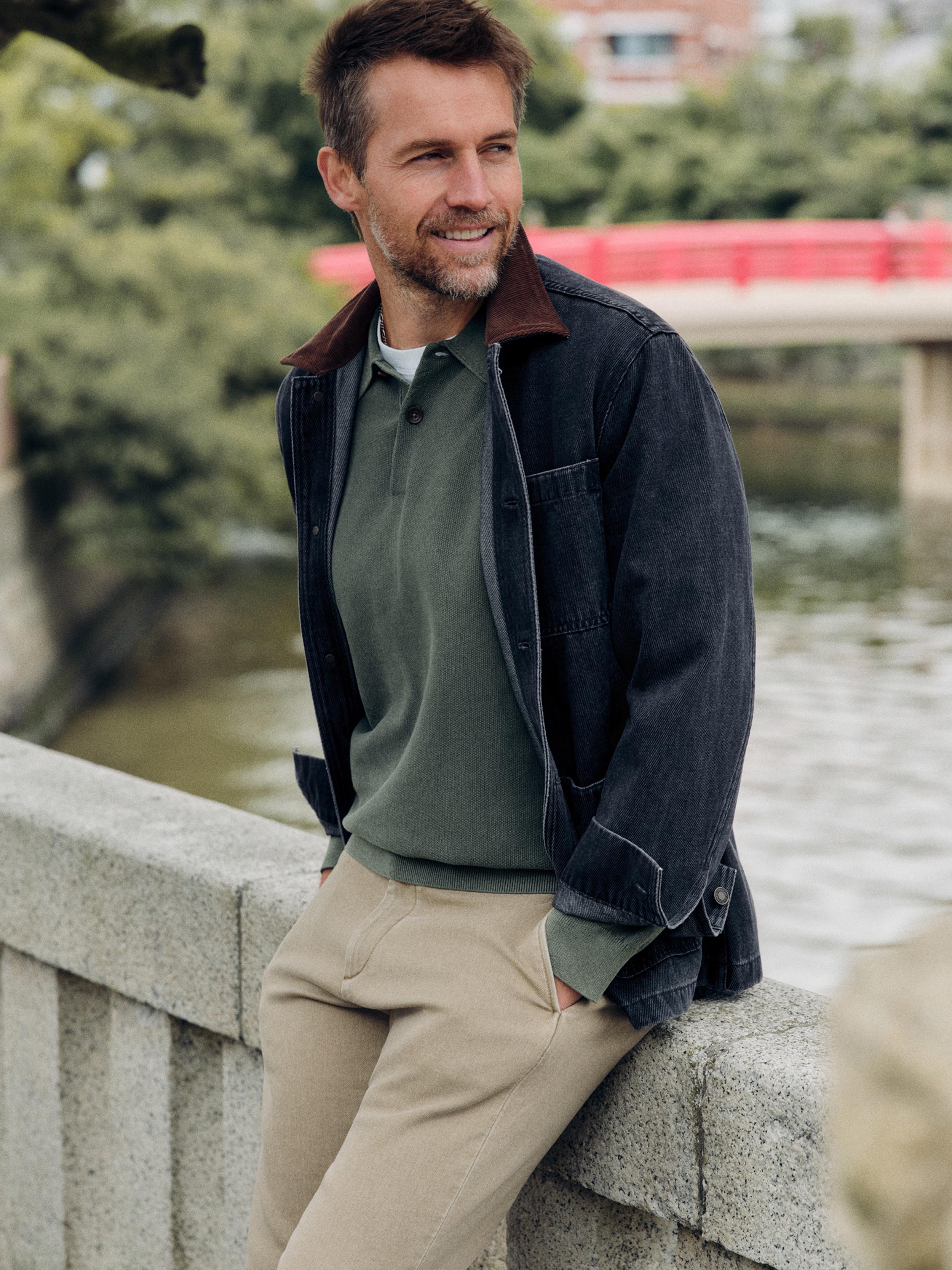A man wearing a dark charcoal corduroy overshirt with a brown collar, an olive green polo shirt, and beige trousers, leaning against a stone bridge railing with water and a red bridge in the background.