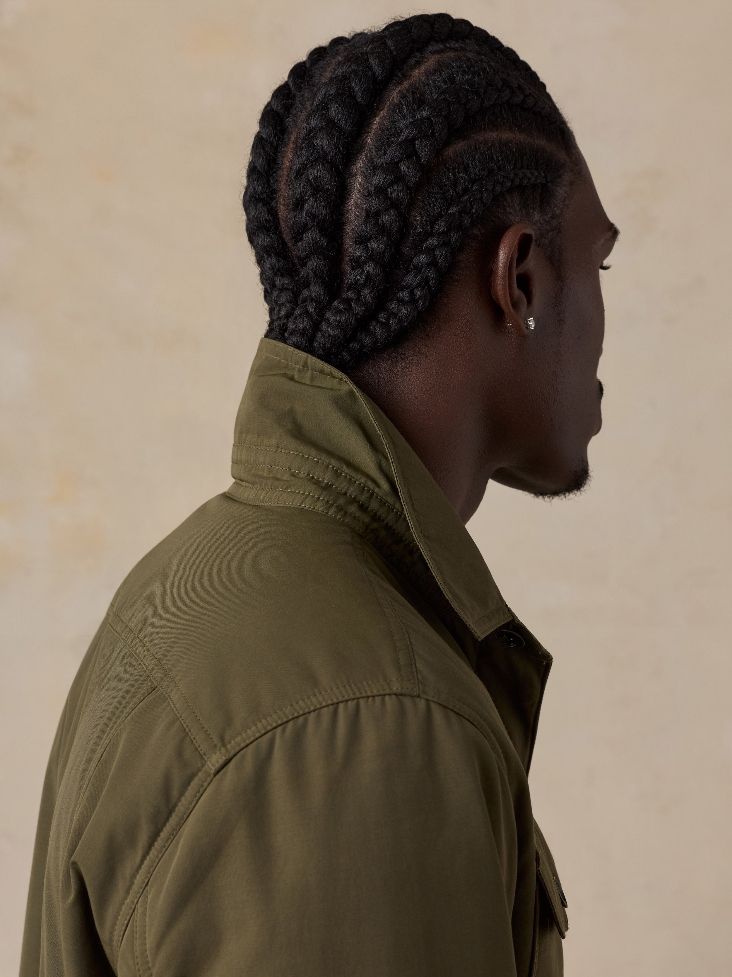 A man shown in profile dressed in an olive green jacket against a textured beige background.