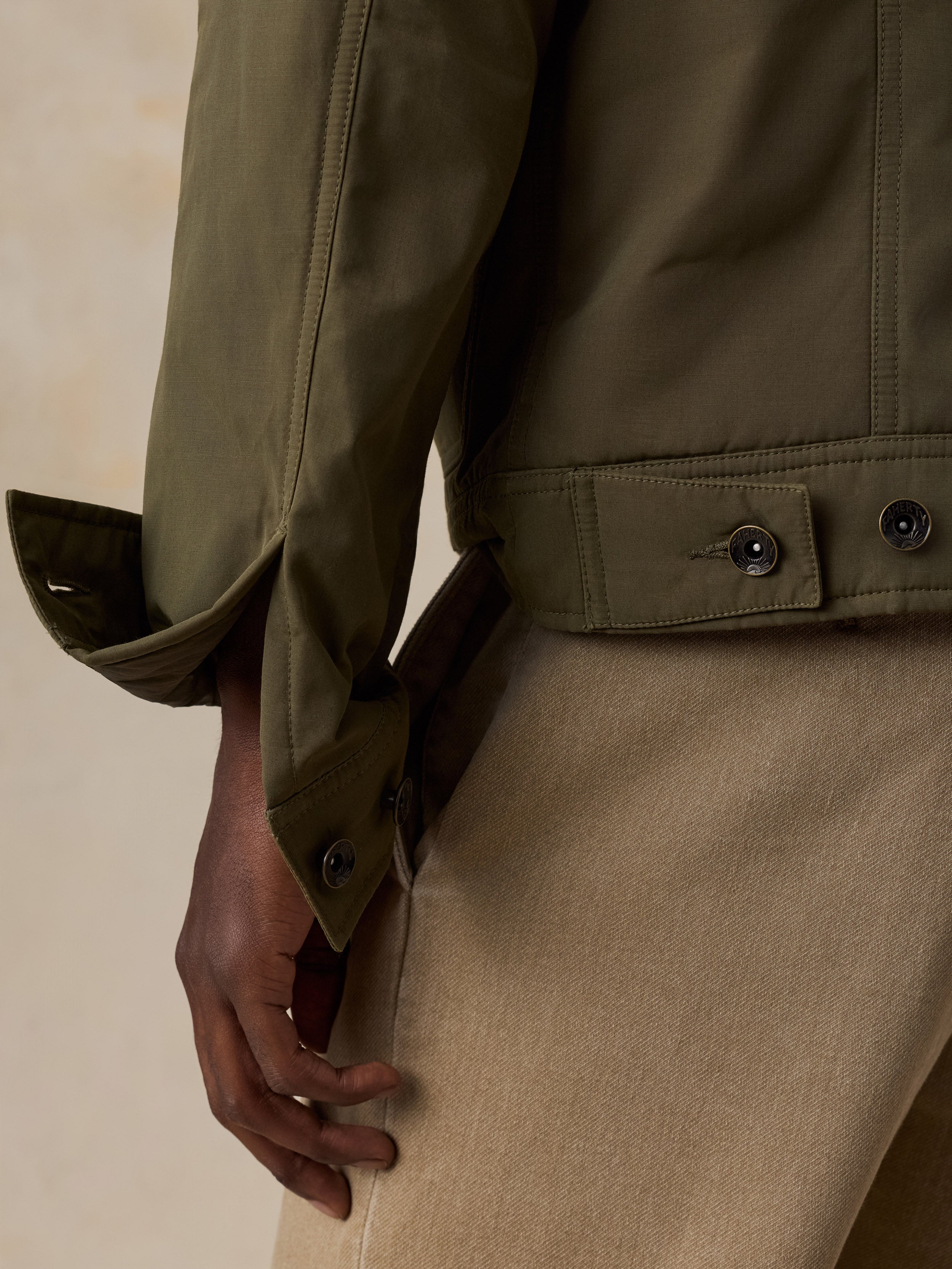 Close-up of a man wearing an olive green jacket with buttoned cuffs and a beige corduroy skirt against a warm beige background.
