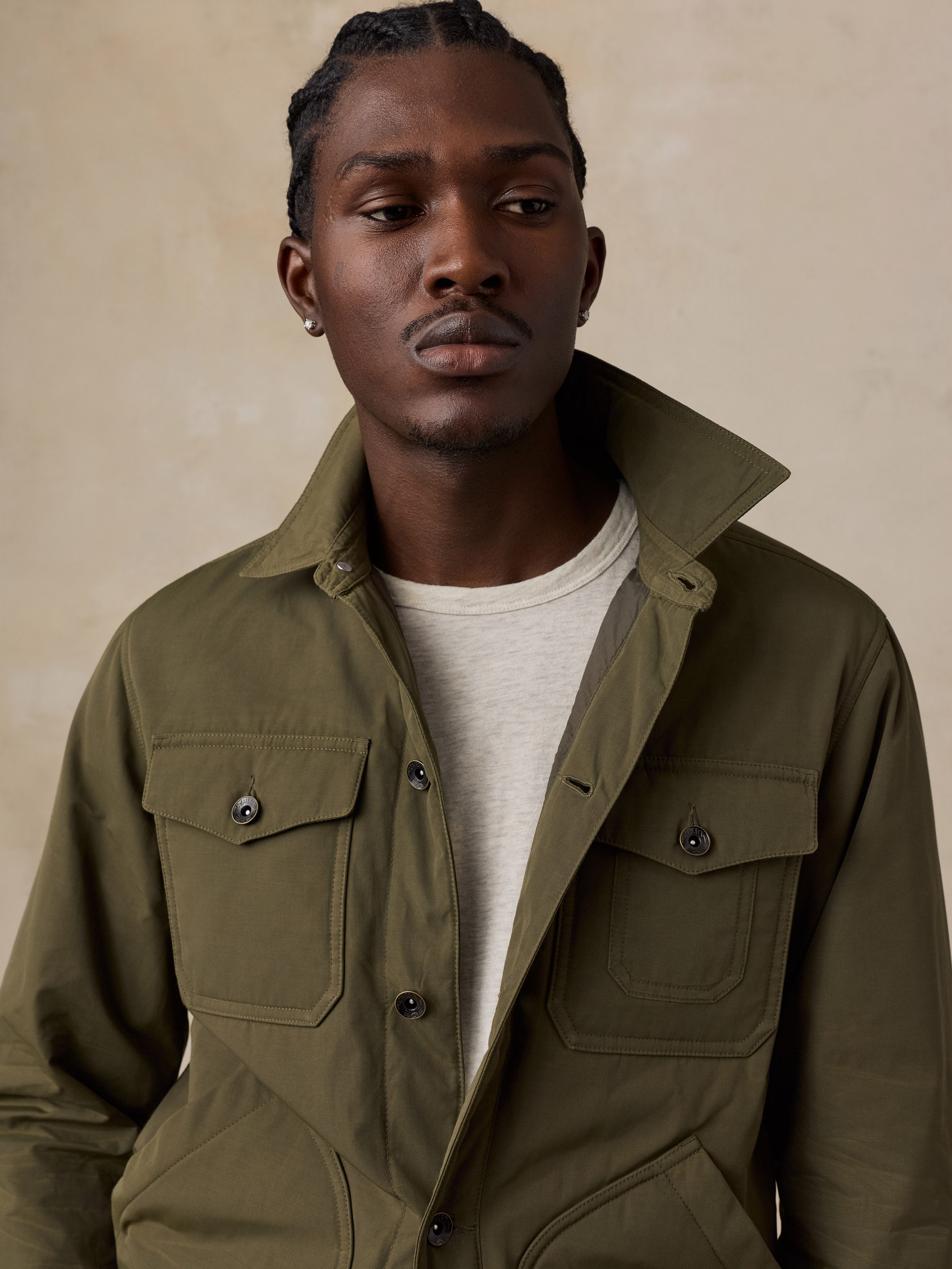A man wearing an olive green utility jacket over a light beige t-shirt, photographed against a textured beige background.