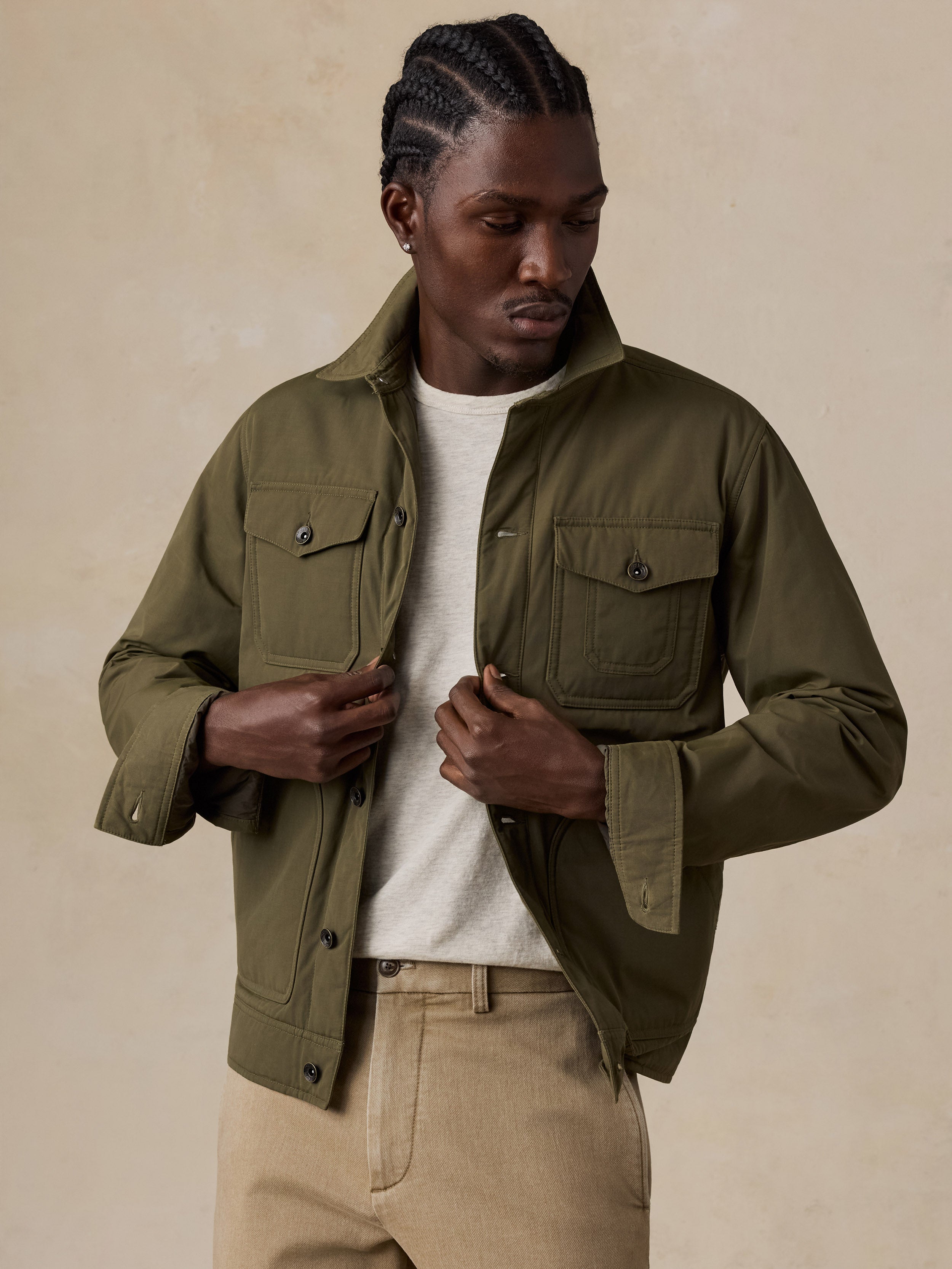 A man wearing an olive green utility jacket over a white t-shirt and beige trousers, standing against a textured beige background.
