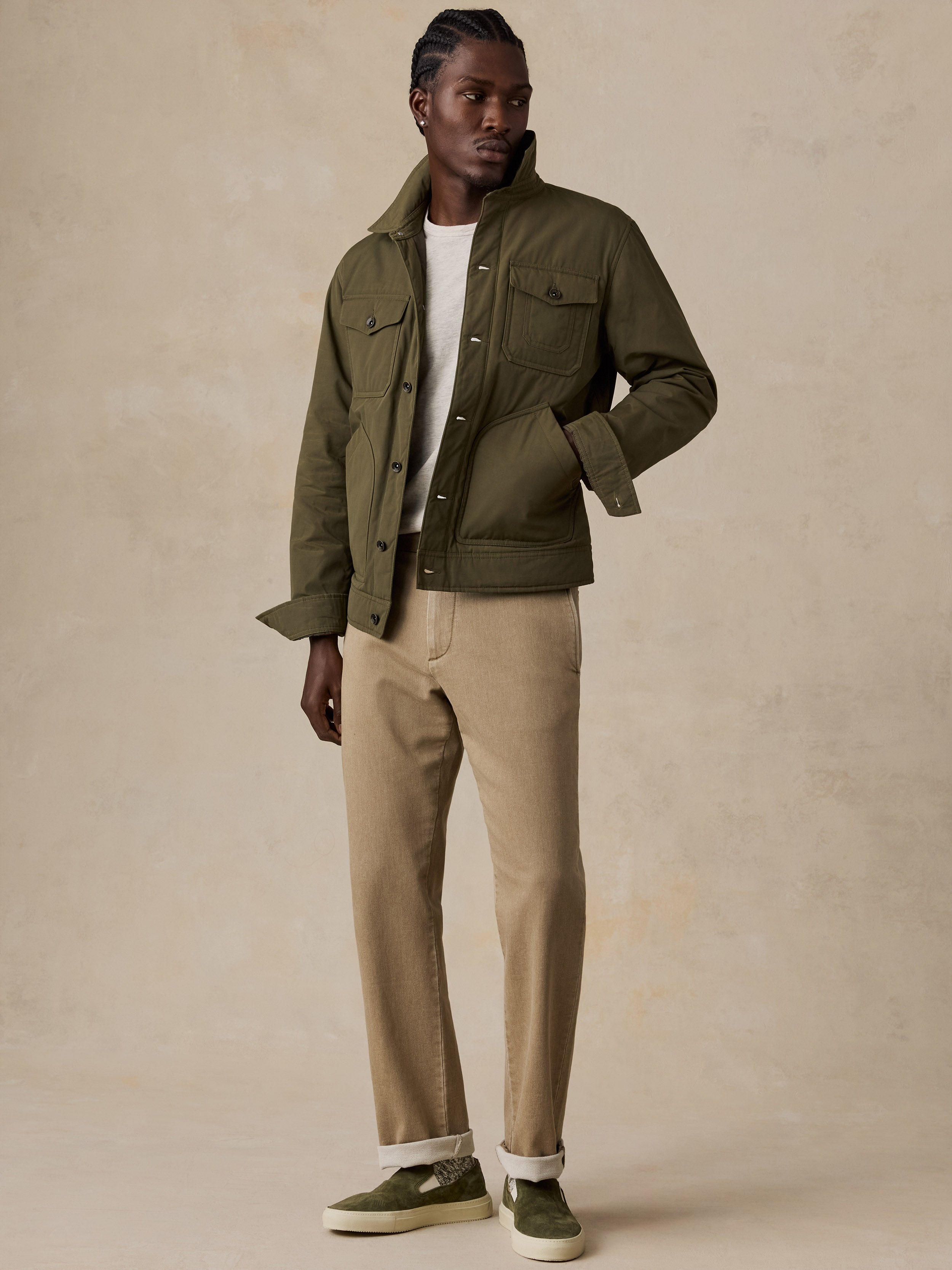 A male model stands against a textured beige studio backdrop wearing an olive green utility jacket over a white t-shirt, beige chinos, and olive green slip-on sneakers.