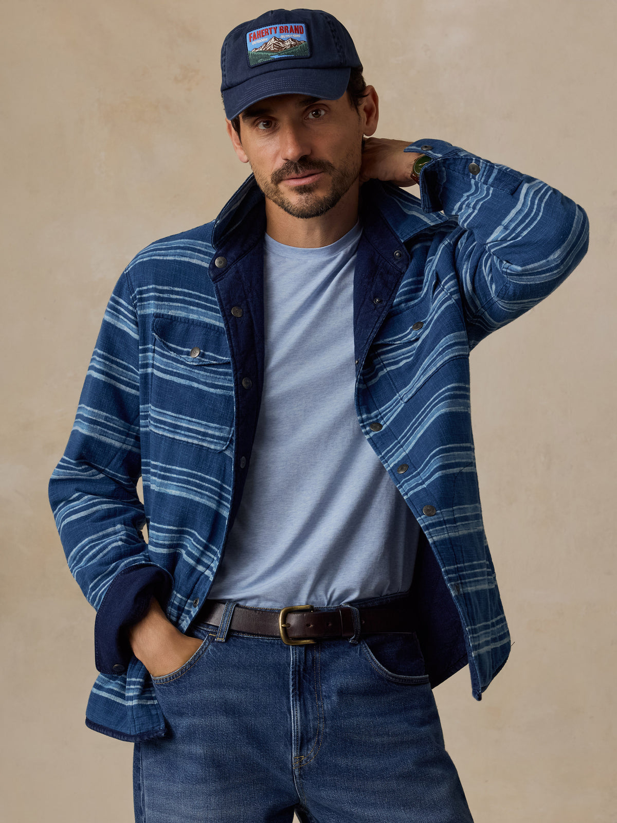A man wearing a blue and navy striped button-up overshirt over a light blue t-shirt, paired with blue jeans and a navy baseball cap with a 'Mountain' patch, posed against a textured beige background.